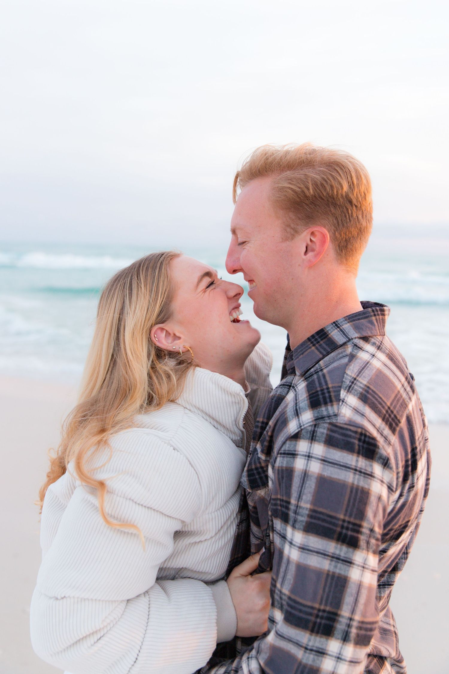 Intimate beach photoshoot featuring couple in casual winter attire against ocean backdrop.