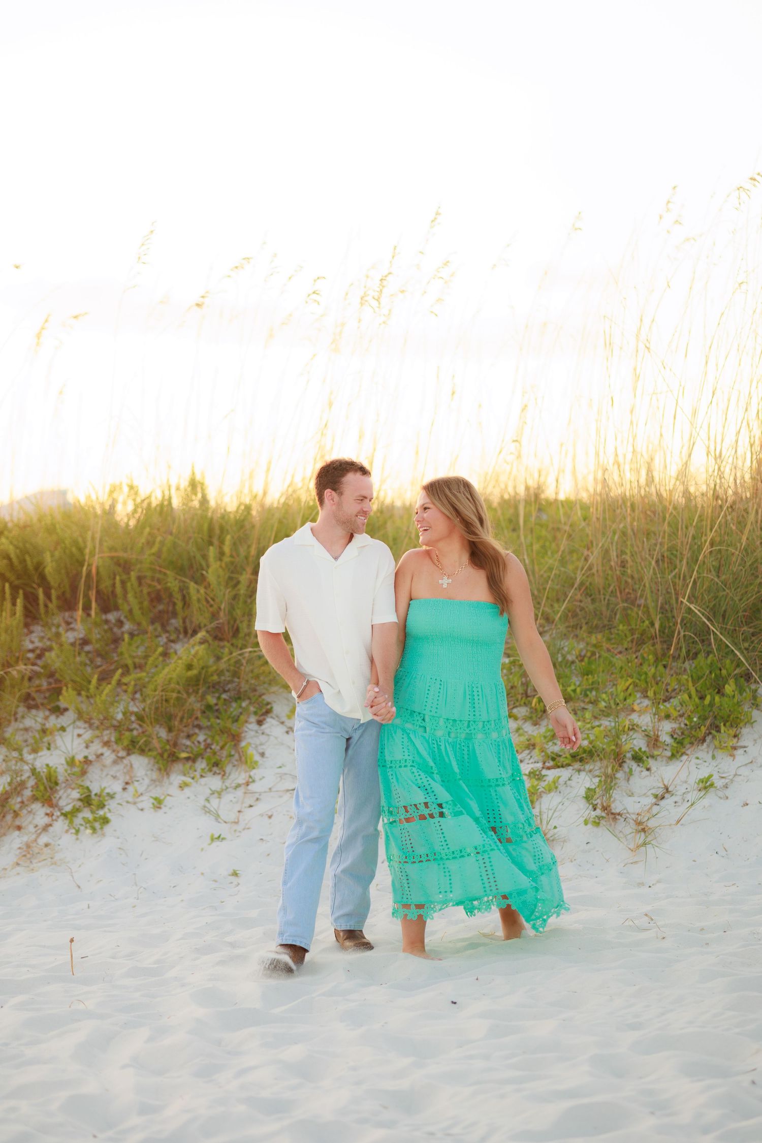 A couple in casual attire walks together on white sand beach dunes during golden hour sunset.