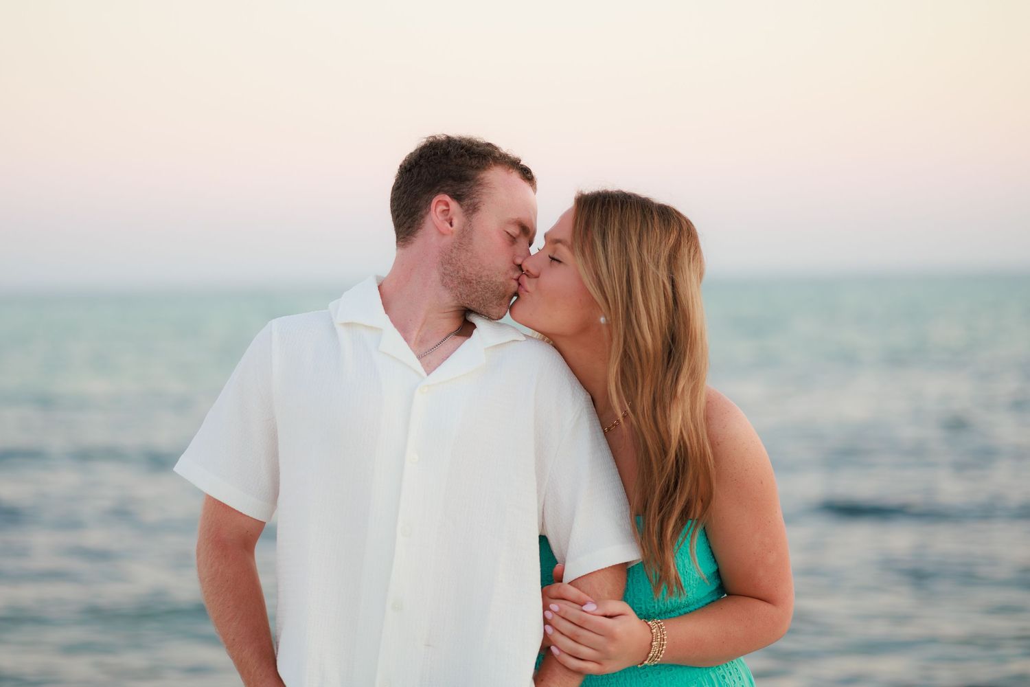 A couple in white and turquoise beachwear embraces on the shore with the ocean in the background at sunset.