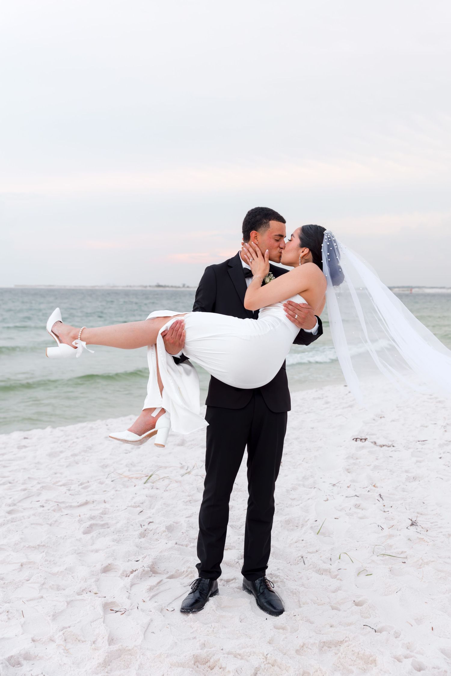 Groom in black suit carries bride in white dress on sandy beach.