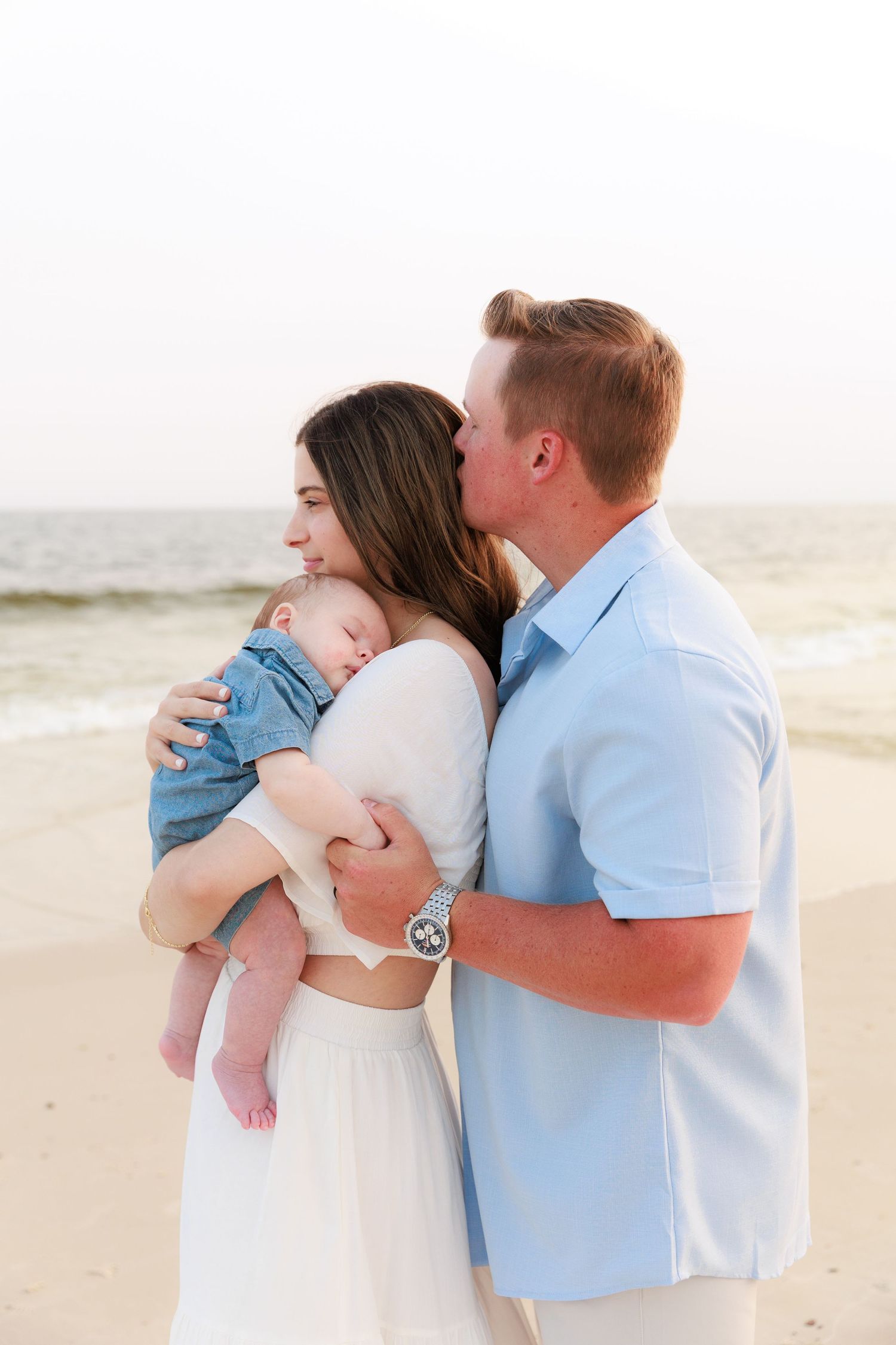 A couple stands on a beach, man kissing woman's head as she holds their sleeping baby dressed in denim.