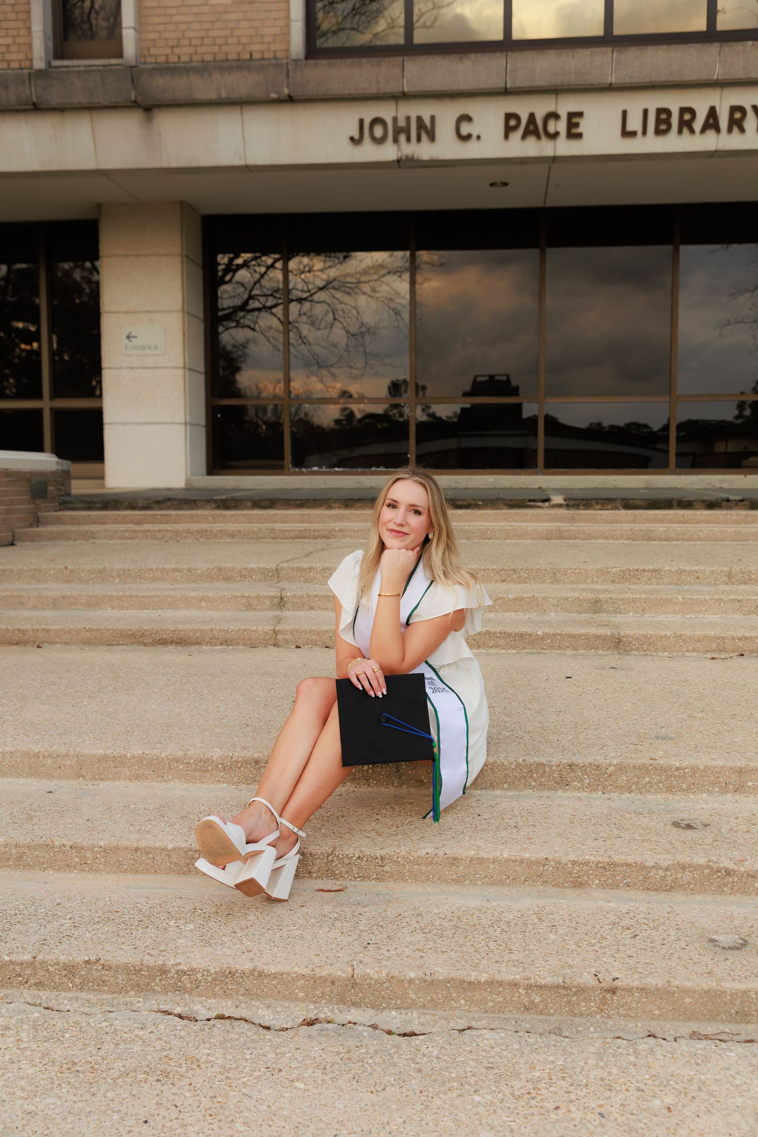 A smiling graduate sits on steps outside John C. Pace Library holding a graduation cap.