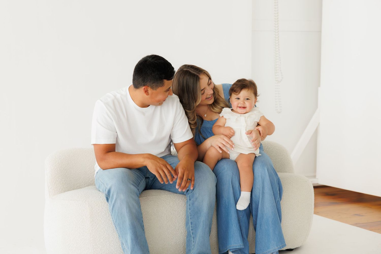 Happy young family with baby sitting on white sofa, parents smiling at infant in bright modern living room.