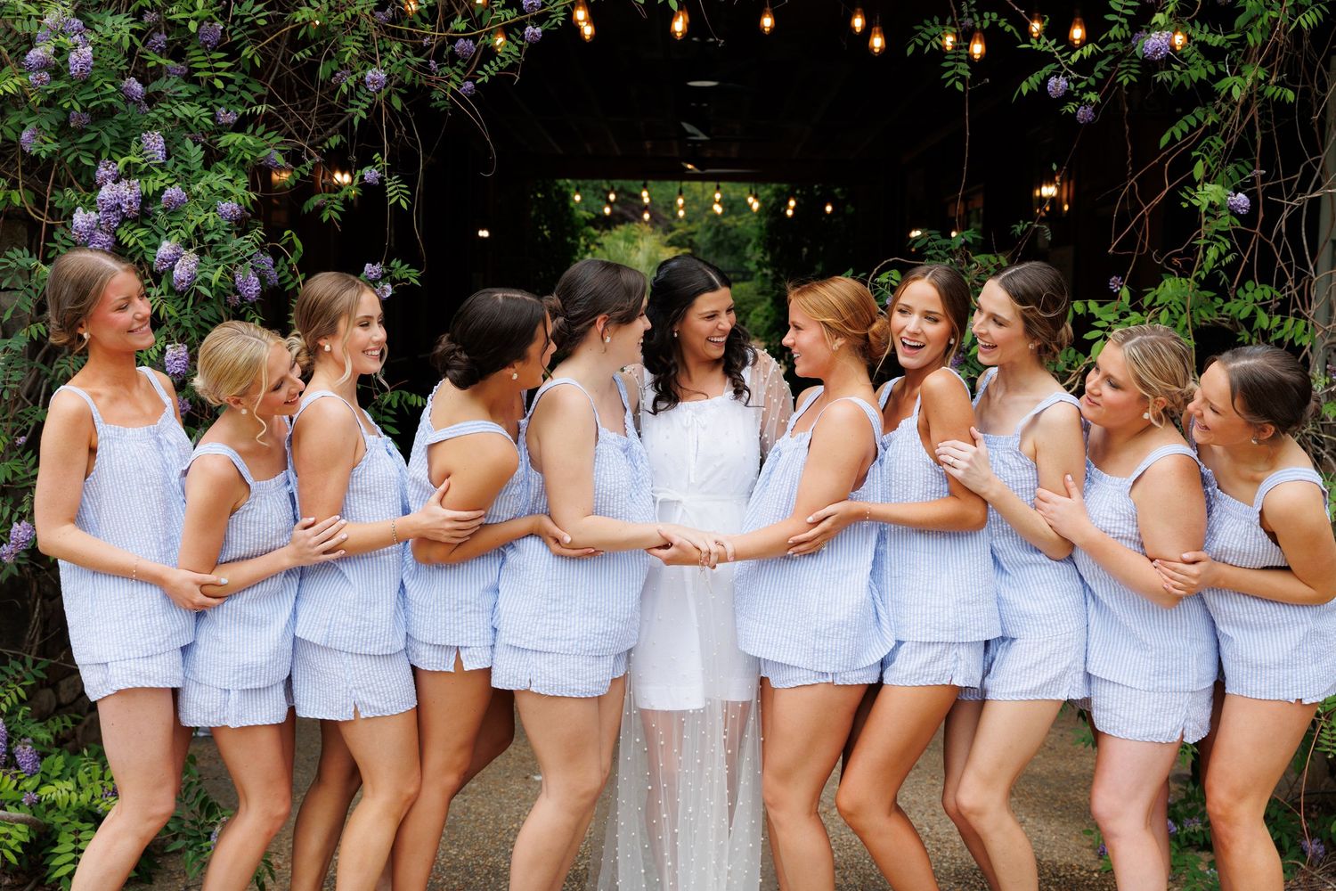 Bride in white gown surrounded by bridesmaids in matching light blue seersucker sets posing under floral arch.