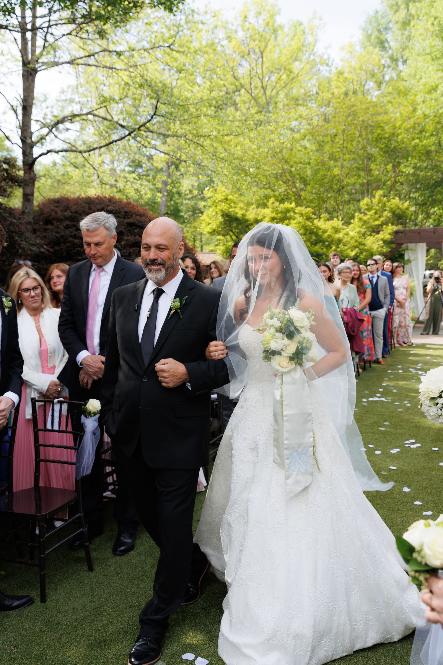 Bride in white gown and veil walks down garden aisle with her father during an elegant outdoor wedding ceremony.