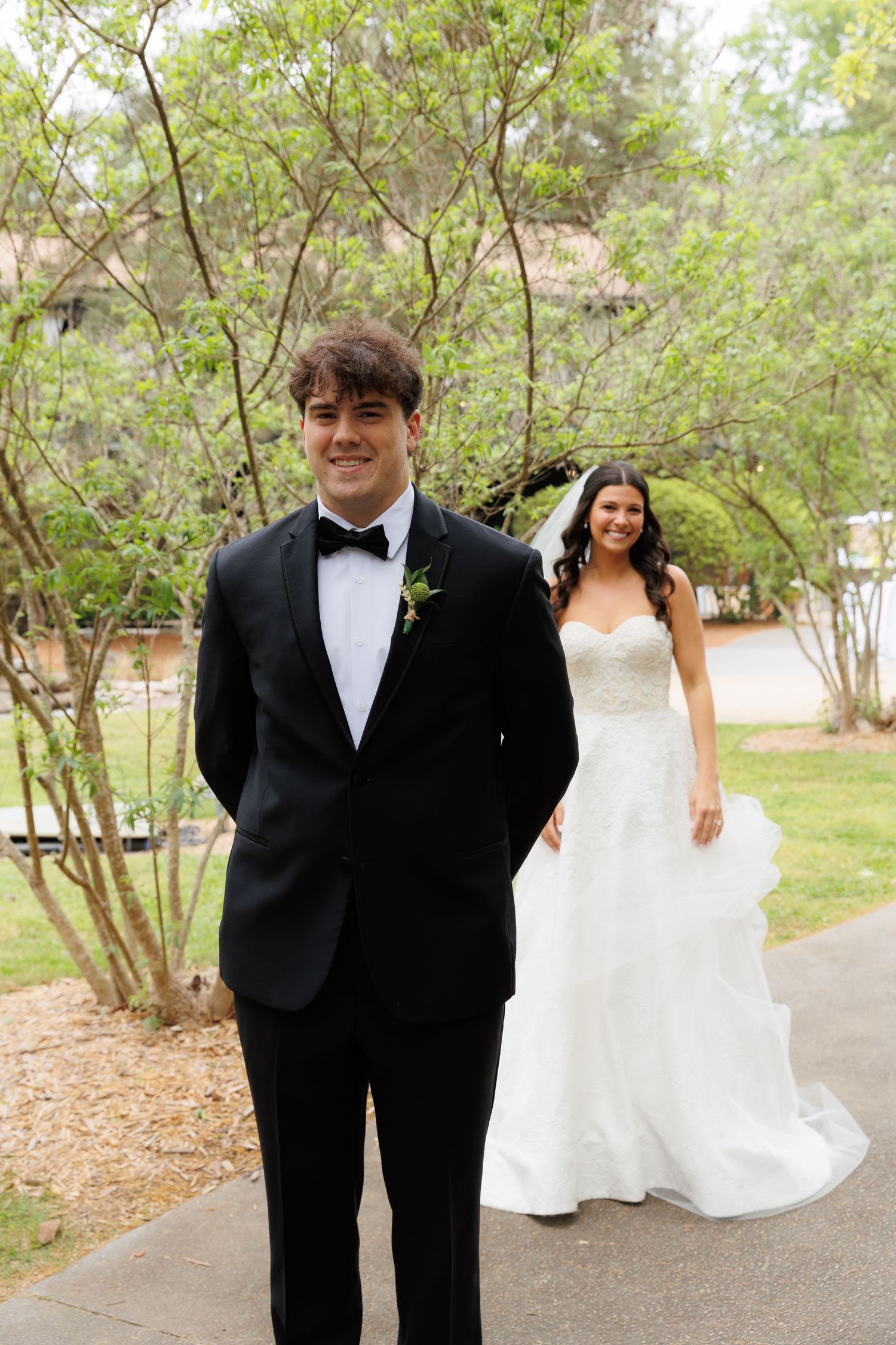 Groom in black tuxedo stands outdoors as bride in white gown approaches for a first look reveal.