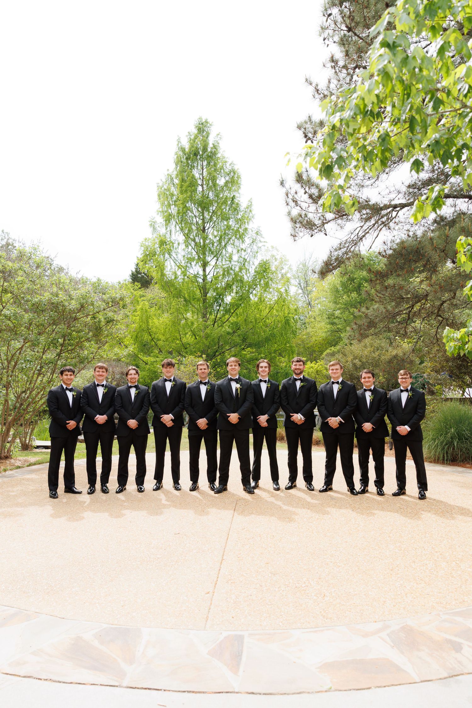 Groom and groomsmen in black tuxedos with bow ties stand in a line outdoors at a wedding venue.