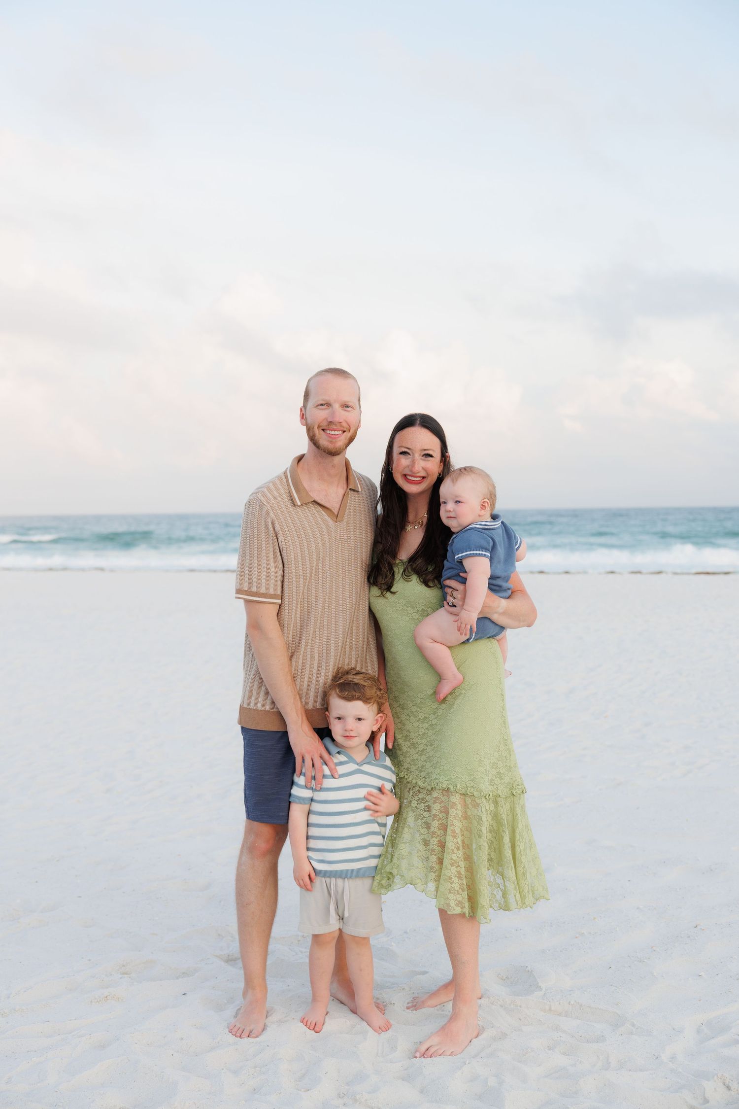 Family of four poses on white sand beach at sunset, parents smiling with toddler son and baby.
