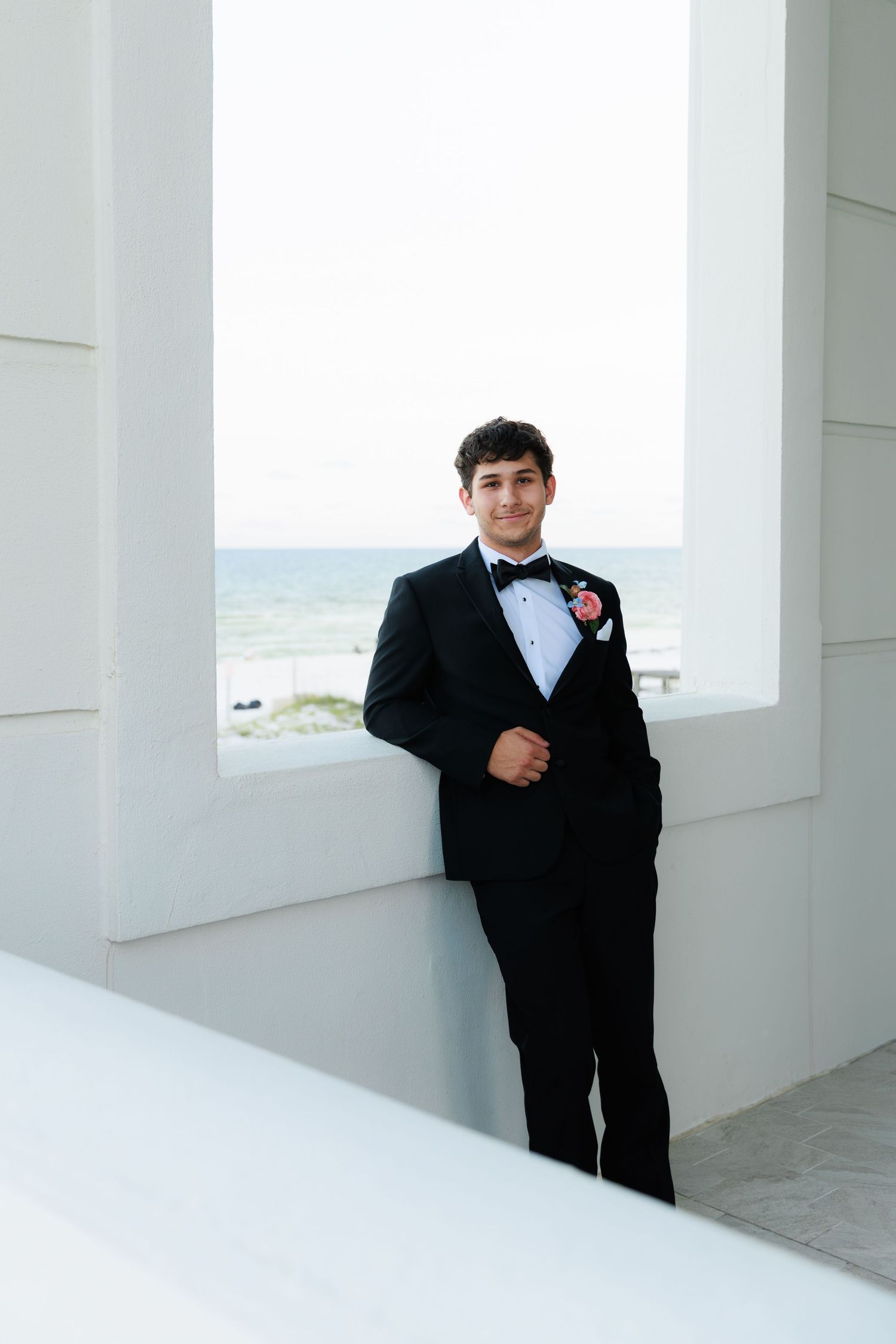 Groom in classic black tuxedo poses against white architectural columns with ocean backdrop.