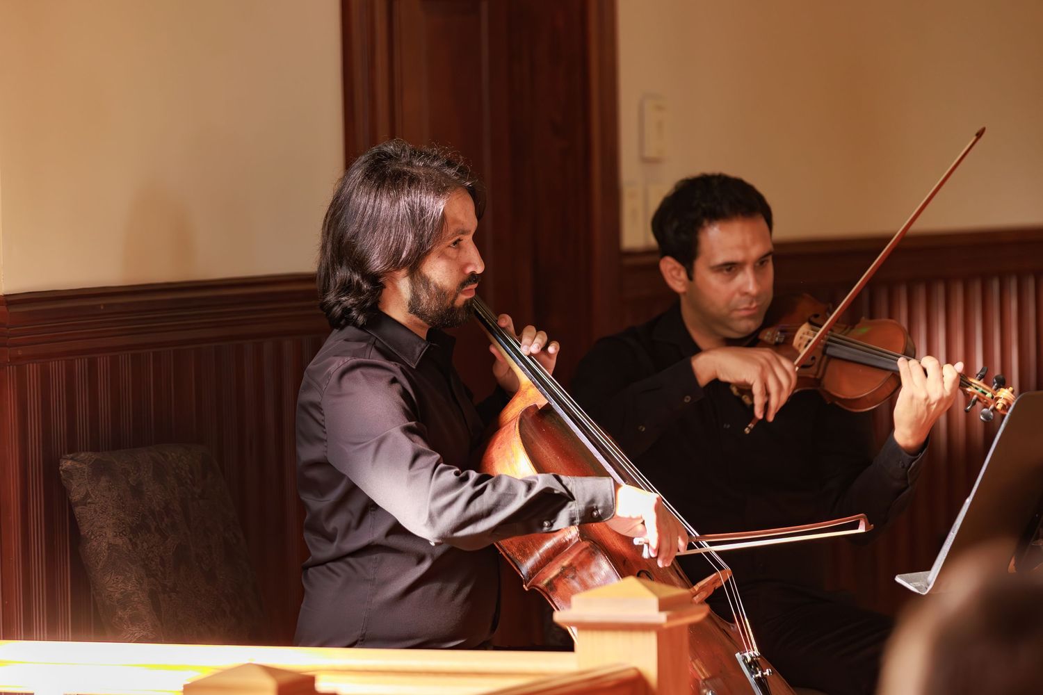 Two musicians performing with string instruments in a wood-paneled venue.