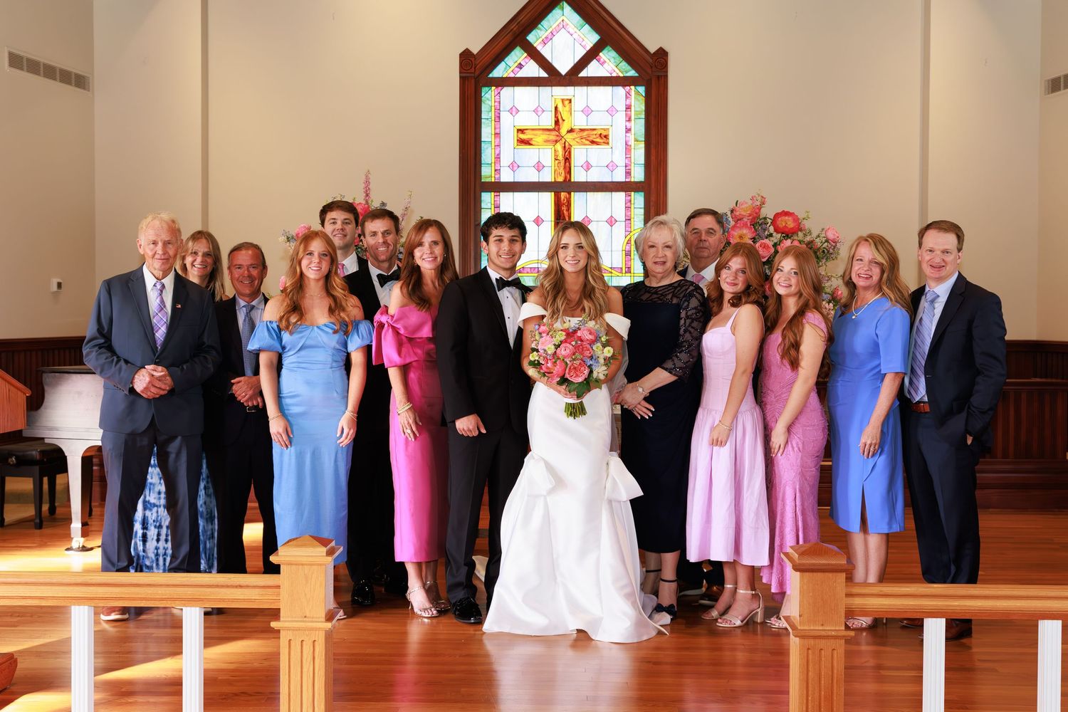 Wedding party group poses in church sanctuary with stained glass window and wooden pews.