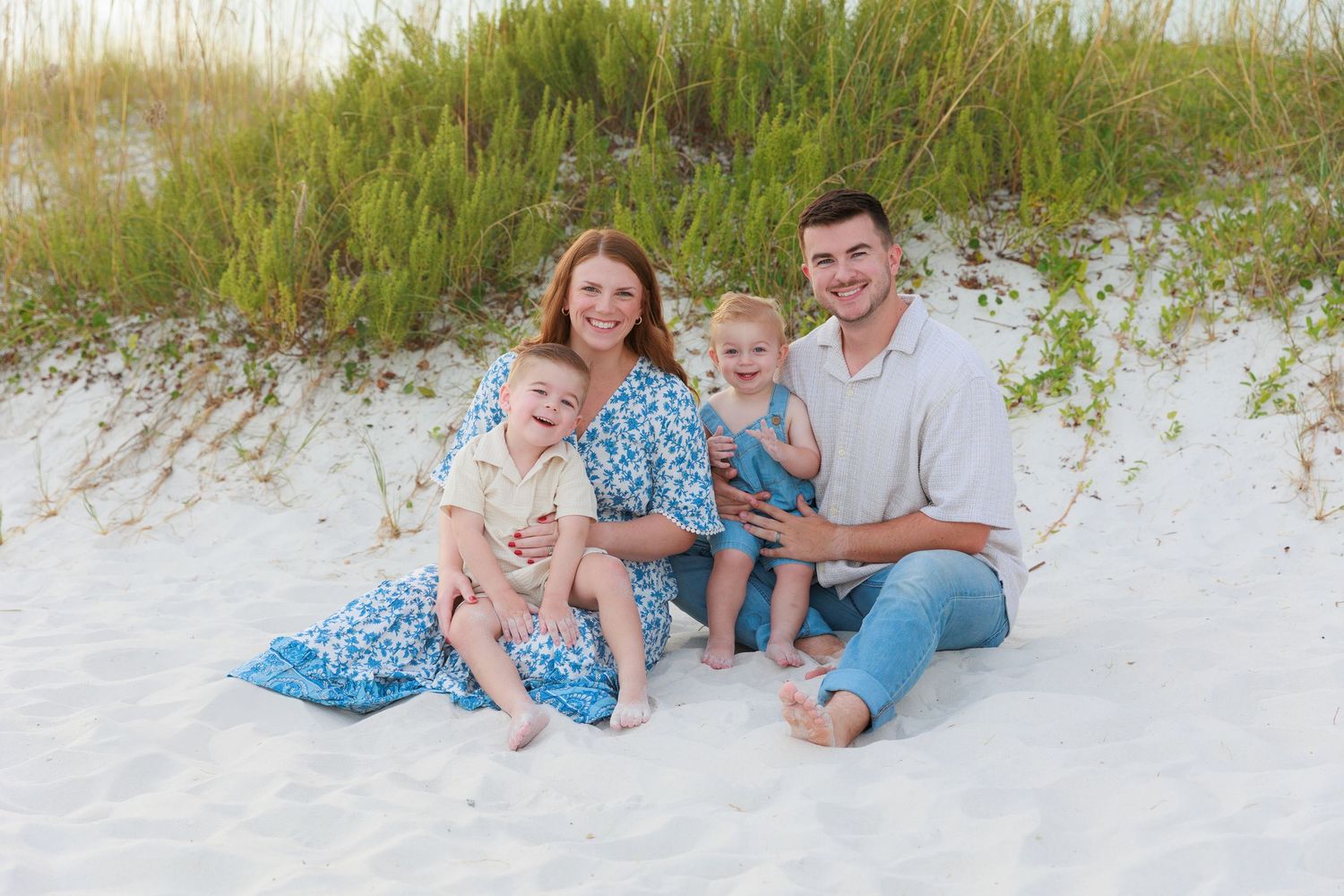 Family of four smiling on white sand beach dunes, parents with two young children in blue and white outfits.