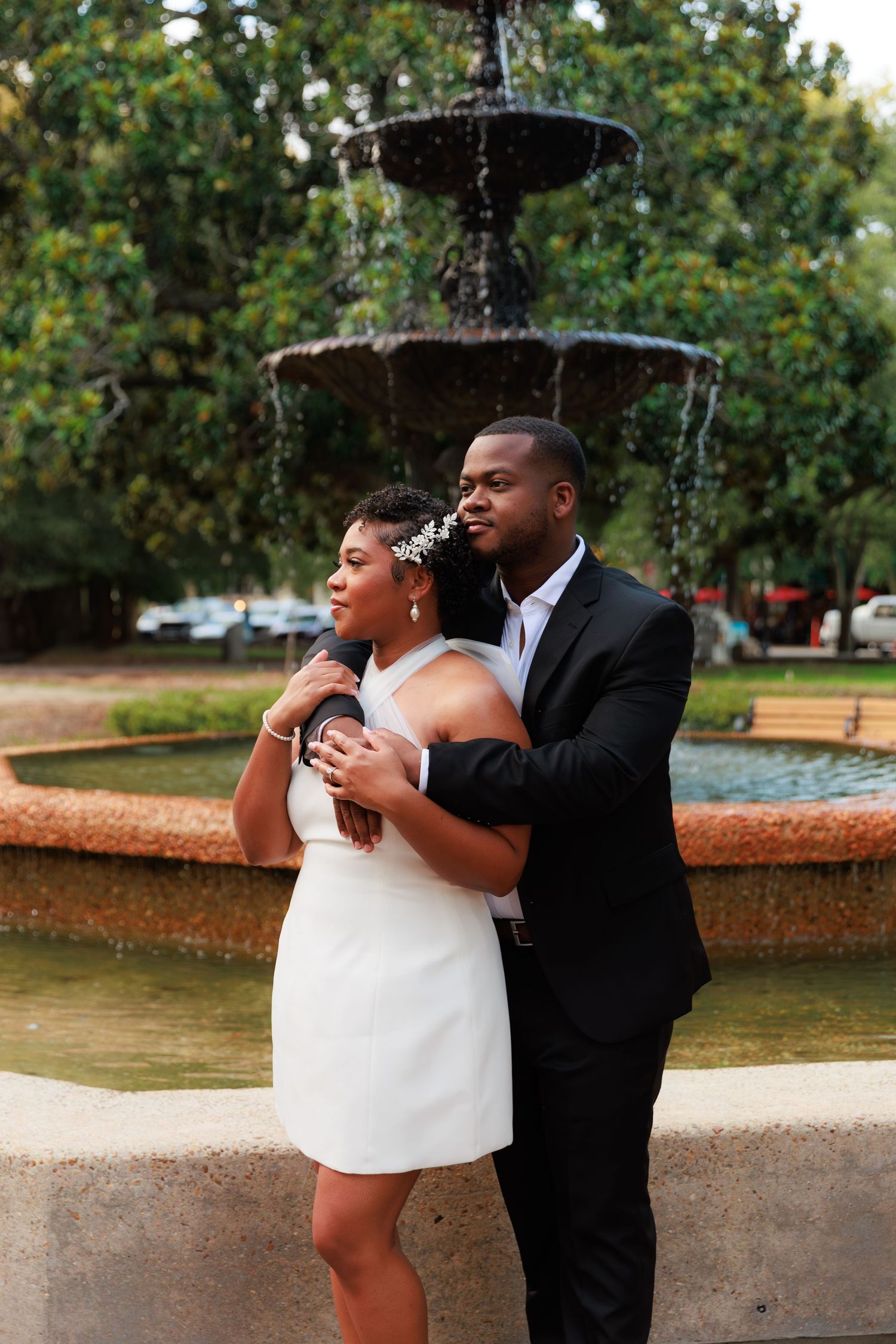 Couple in formal attire embracing by a tiered stone fountain in a lush green park setting.