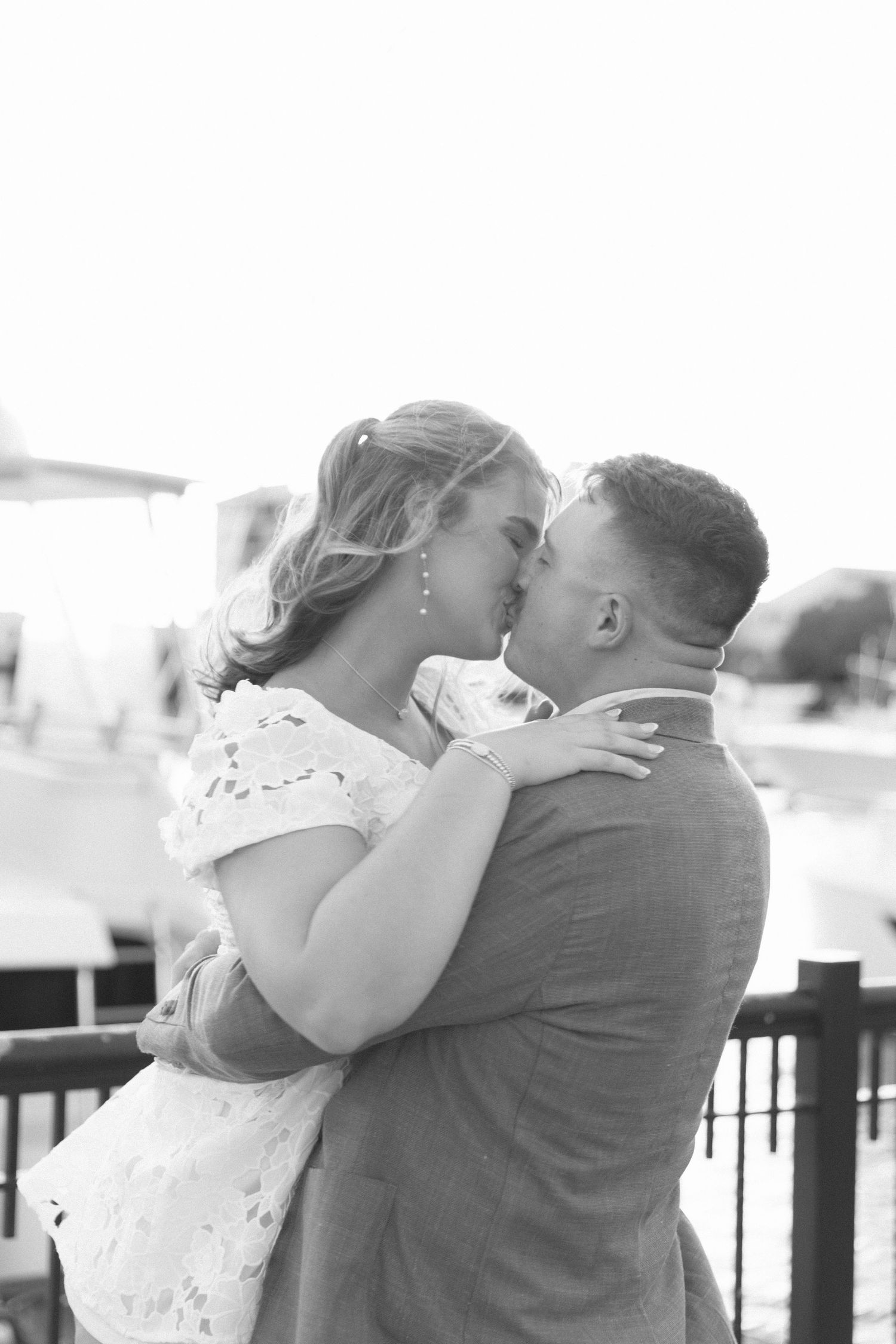 Black and white photo of couple sharing a tender kiss on a waterfront dock, woman in lace dress.