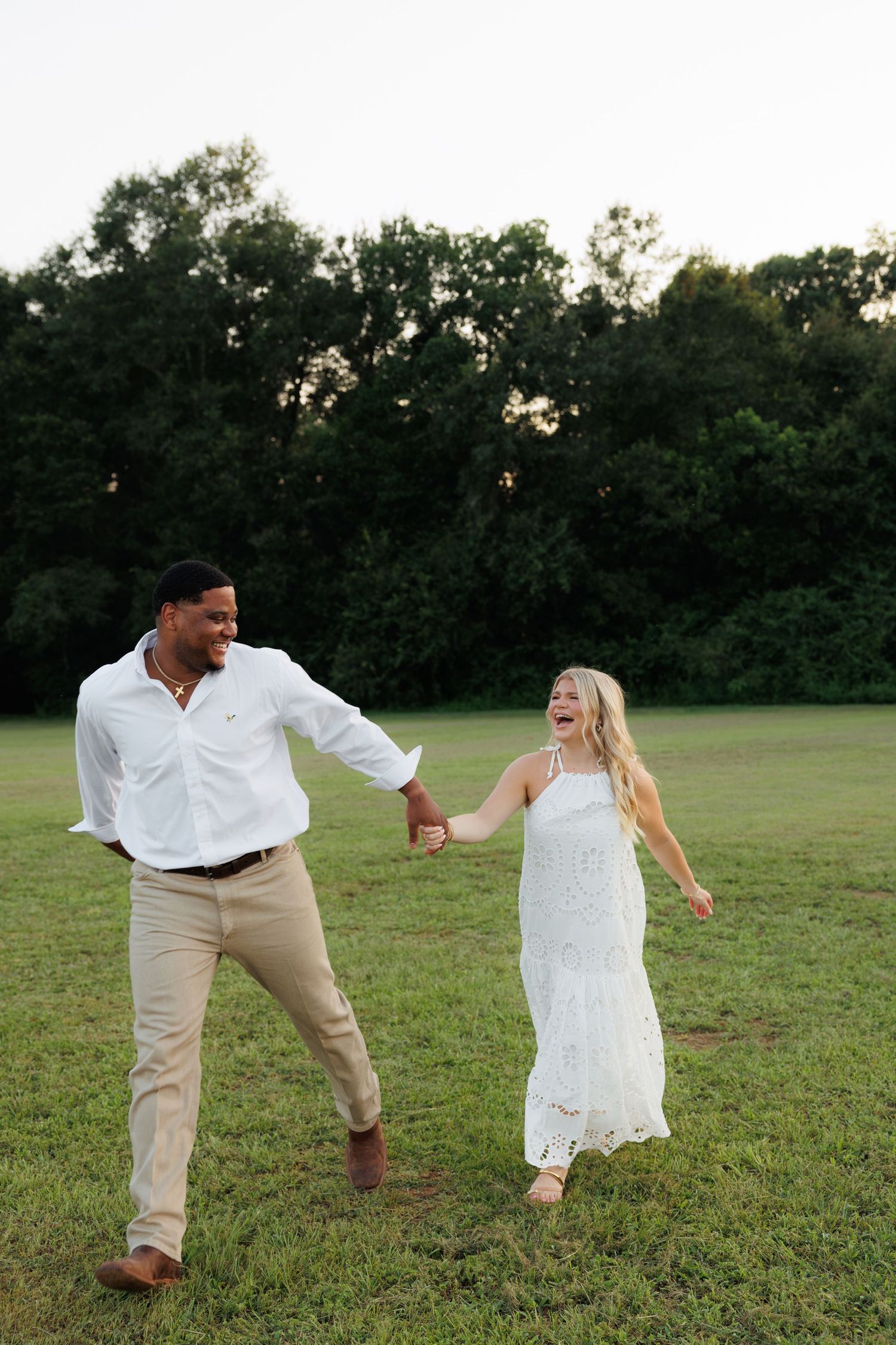 Happy couple in white outfits holding hands and laughing while walking through a lush green field at sunset.
