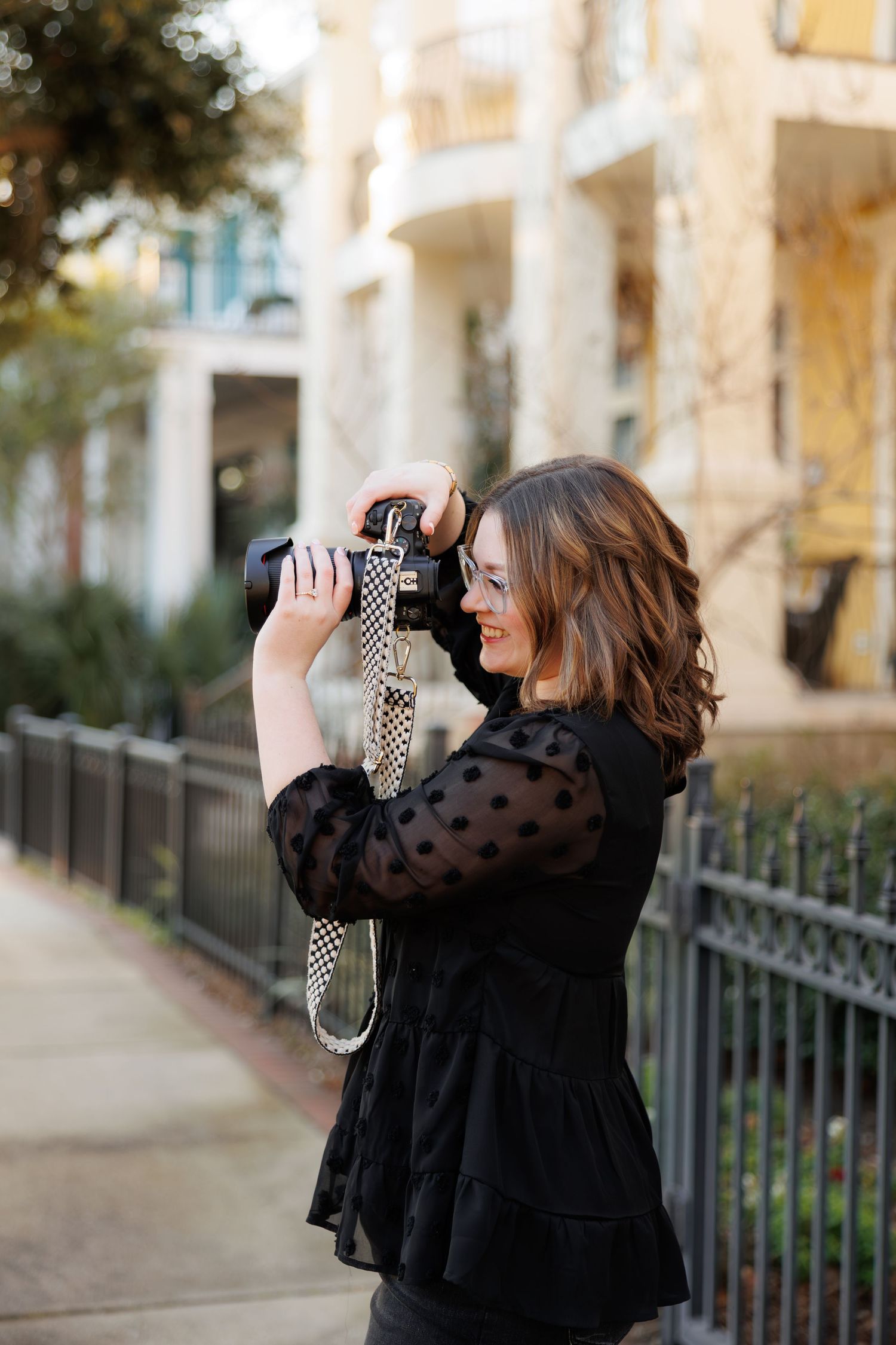 A smiling woman in a black polka dot blouse photographs outdoors with a camera and patterned strap on an urban sidewalk.