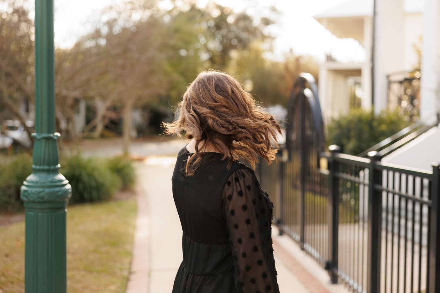 Woman in black polka dot dress with flowing hair walks along a tree-lined sidewalk near an iron fence.