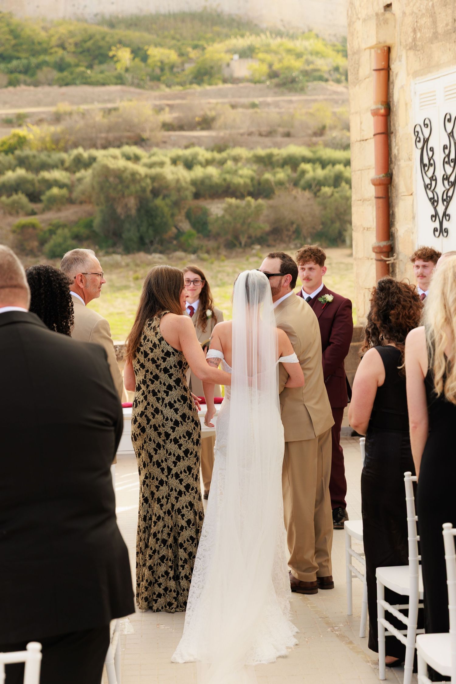 Wedding ceremony guests gather on an outdoor terrace with white chairs and scenic mountain views in the background.