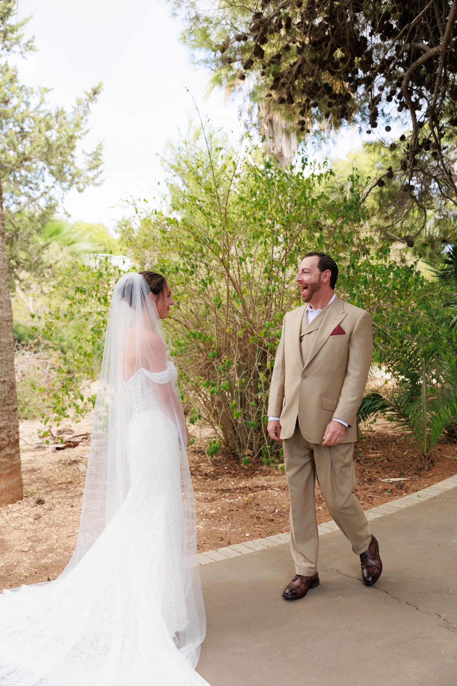 A bride in a flowing white gown and veil stands on a path with someone in a tan suit in a garden setting.