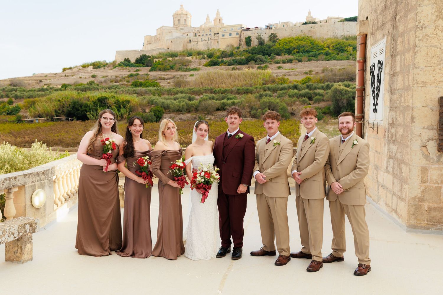 Wedding party in brown and beige attire poses together in front of a rustic stone wall at an outdoor venue.