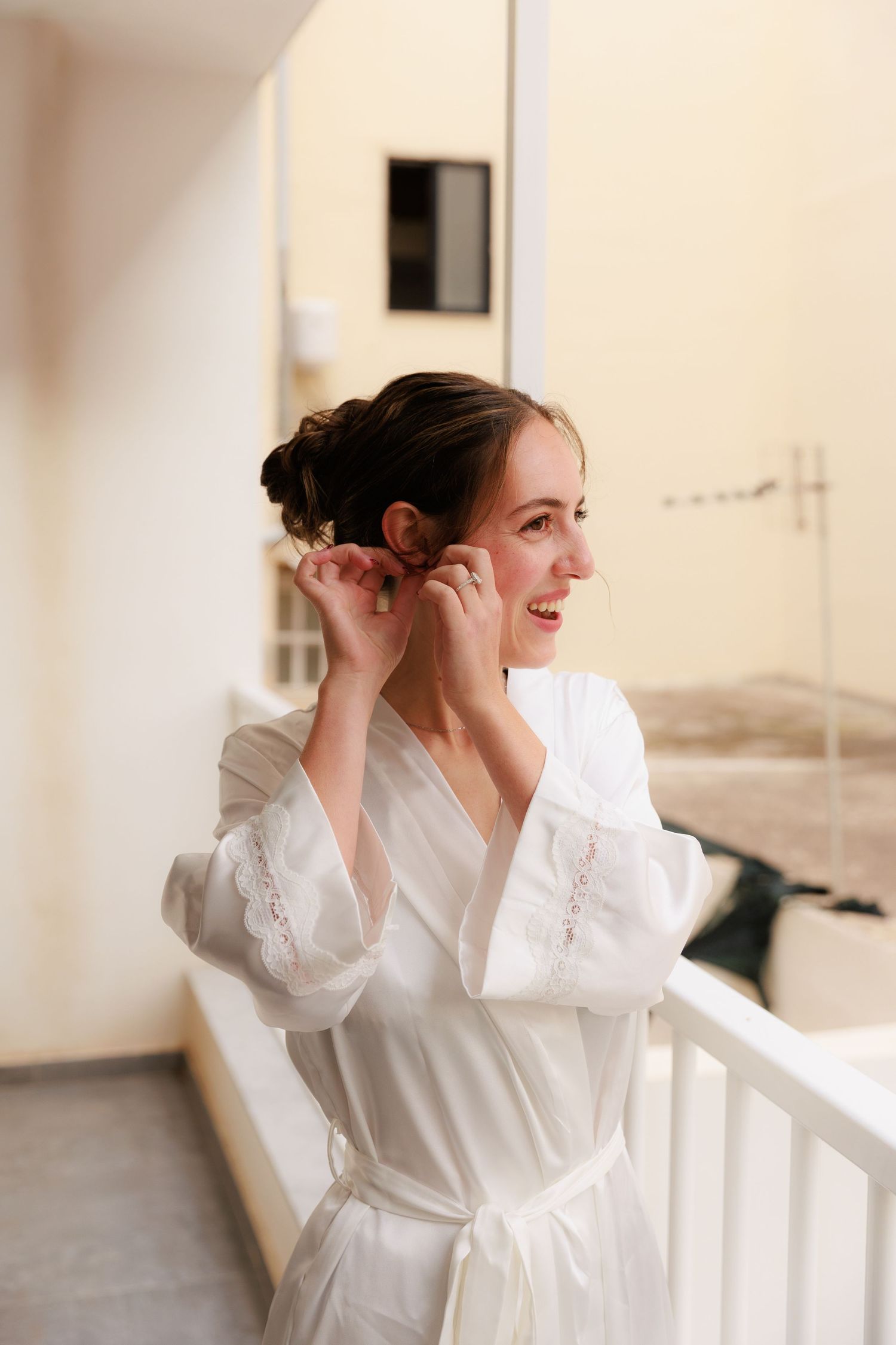 A person in a white silk robe smiles while putting on earrings on a balcony.
