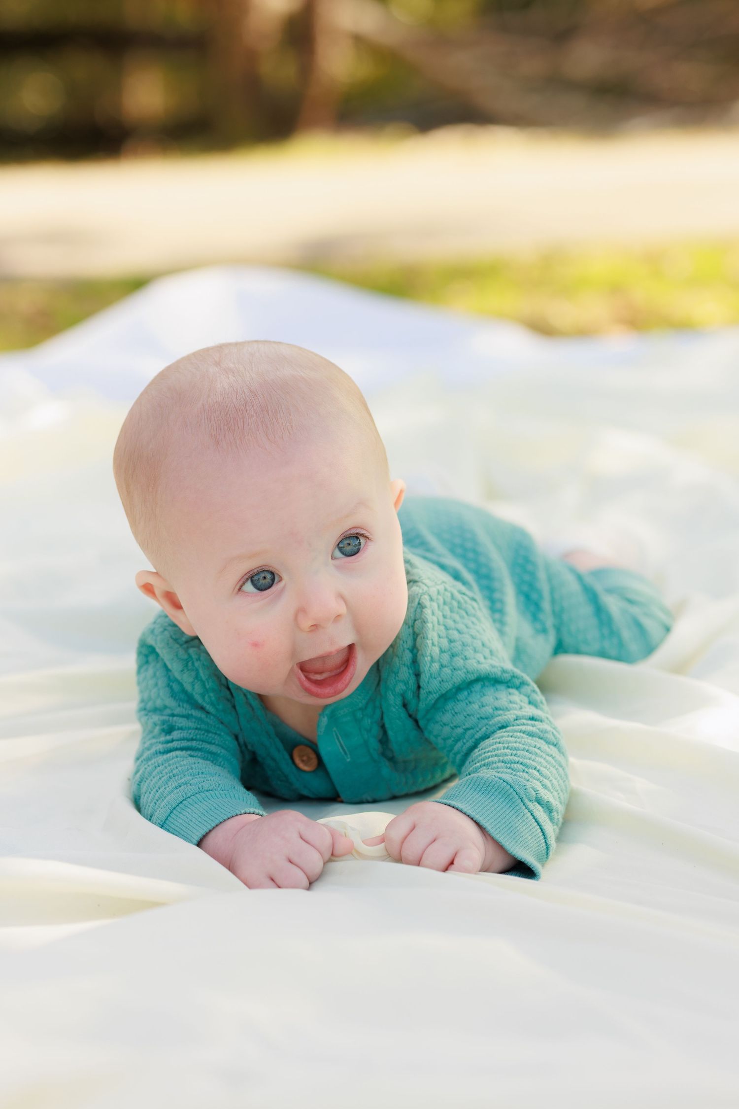Baby in turquoise cardigan smiling while lying on white blanket outdoors.
