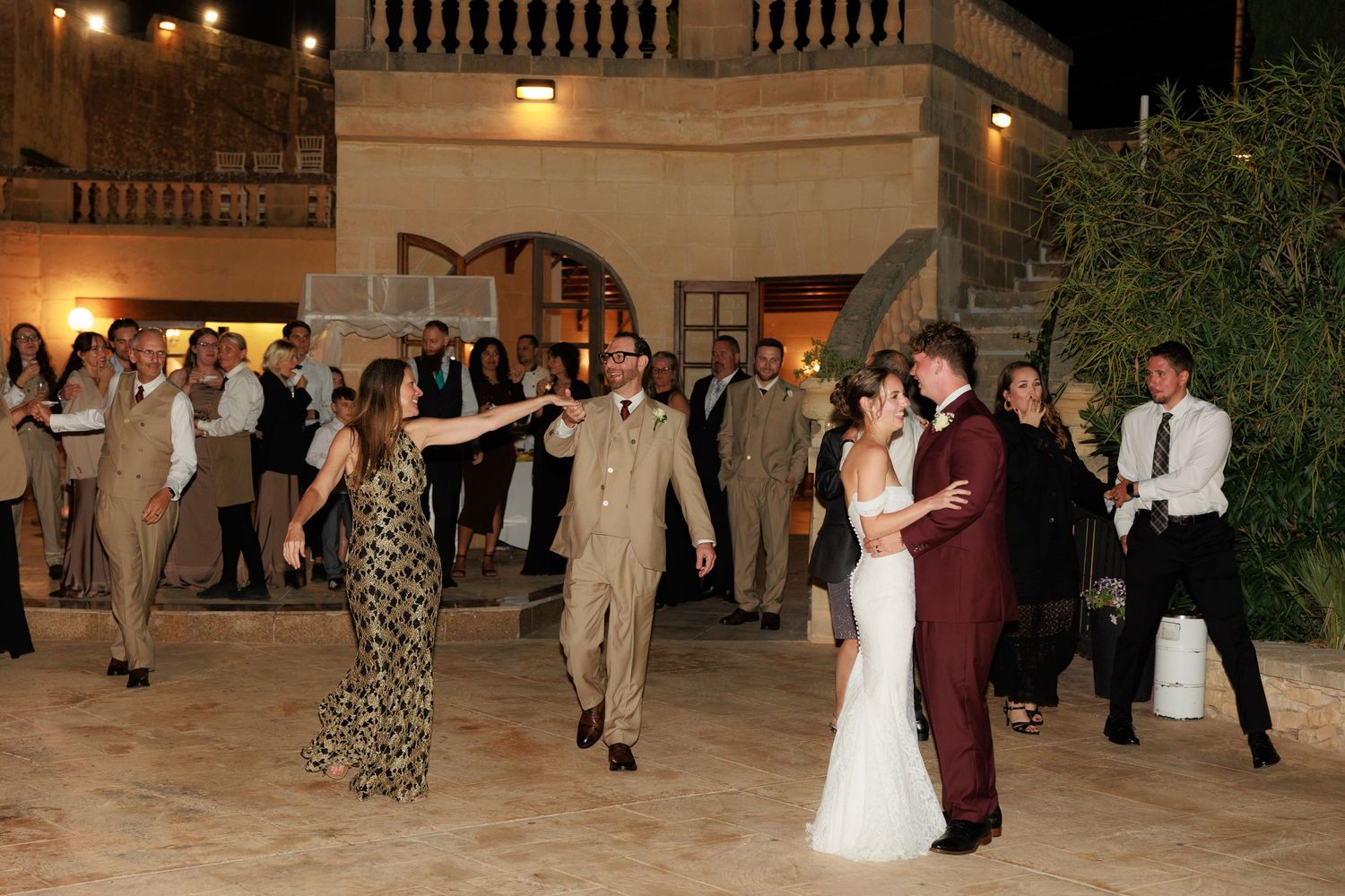 Wedding guests dance together during an evening reception in an elegant ballroom with warm lighting and stone columns.