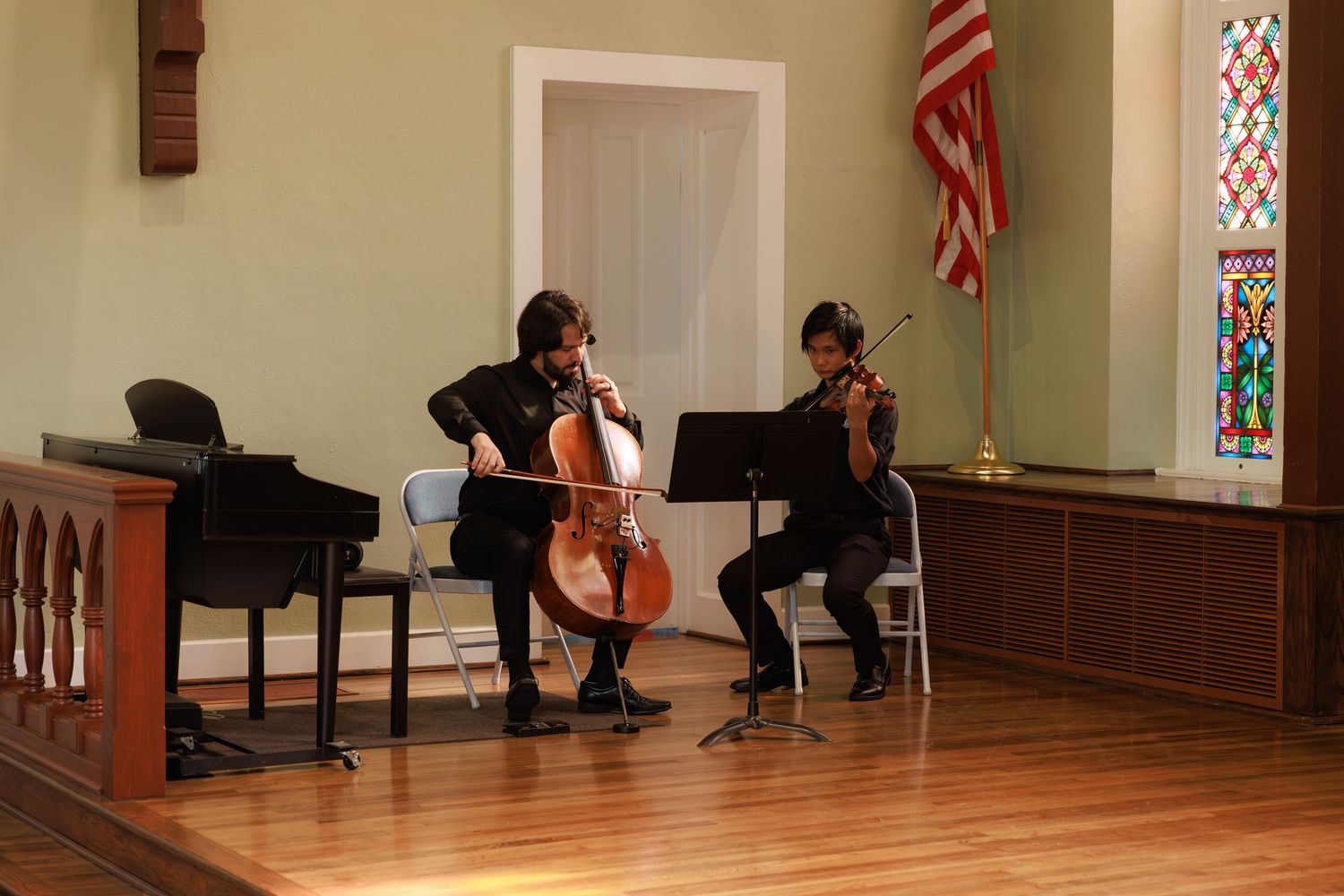 Musicians perform a classical duet with cello and violin in a church sanctuary with wooden floors and stained glass windows.