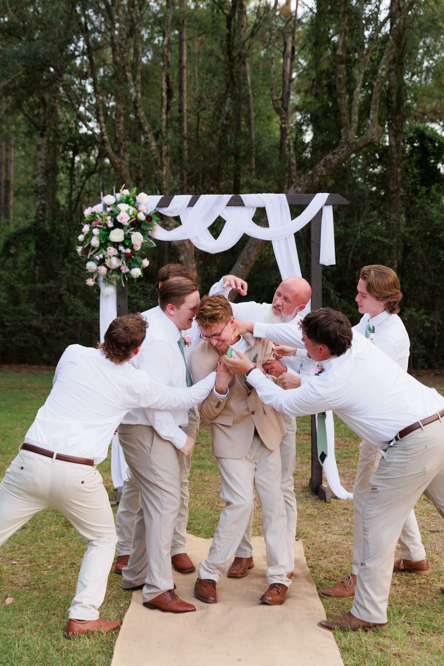 Groomsmen in khaki pants and white shirts playfully pose under a floral wedding arch during outdoor celebration.