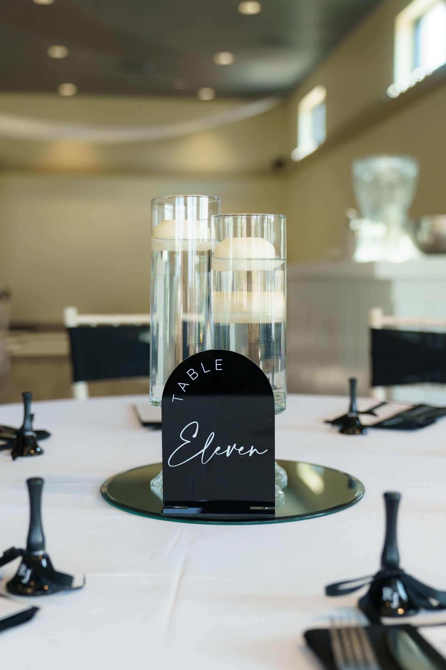Black table number signs with elegant script writing displayed on mirrored stands at an event setting.