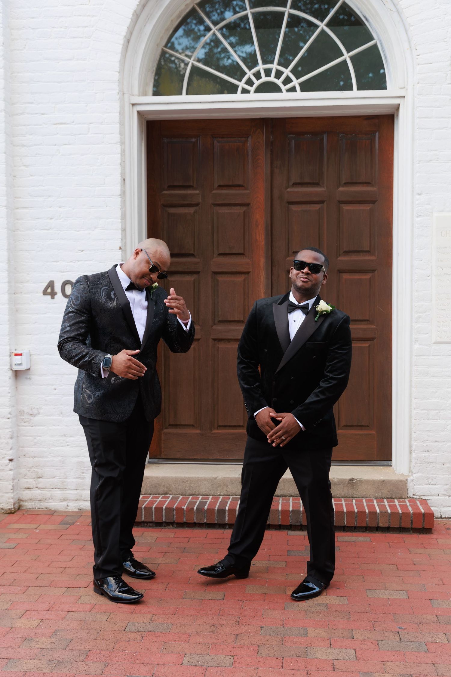 Two groomsmen in black tuxedos and sunglasses pose confidently in front of a brown church door.