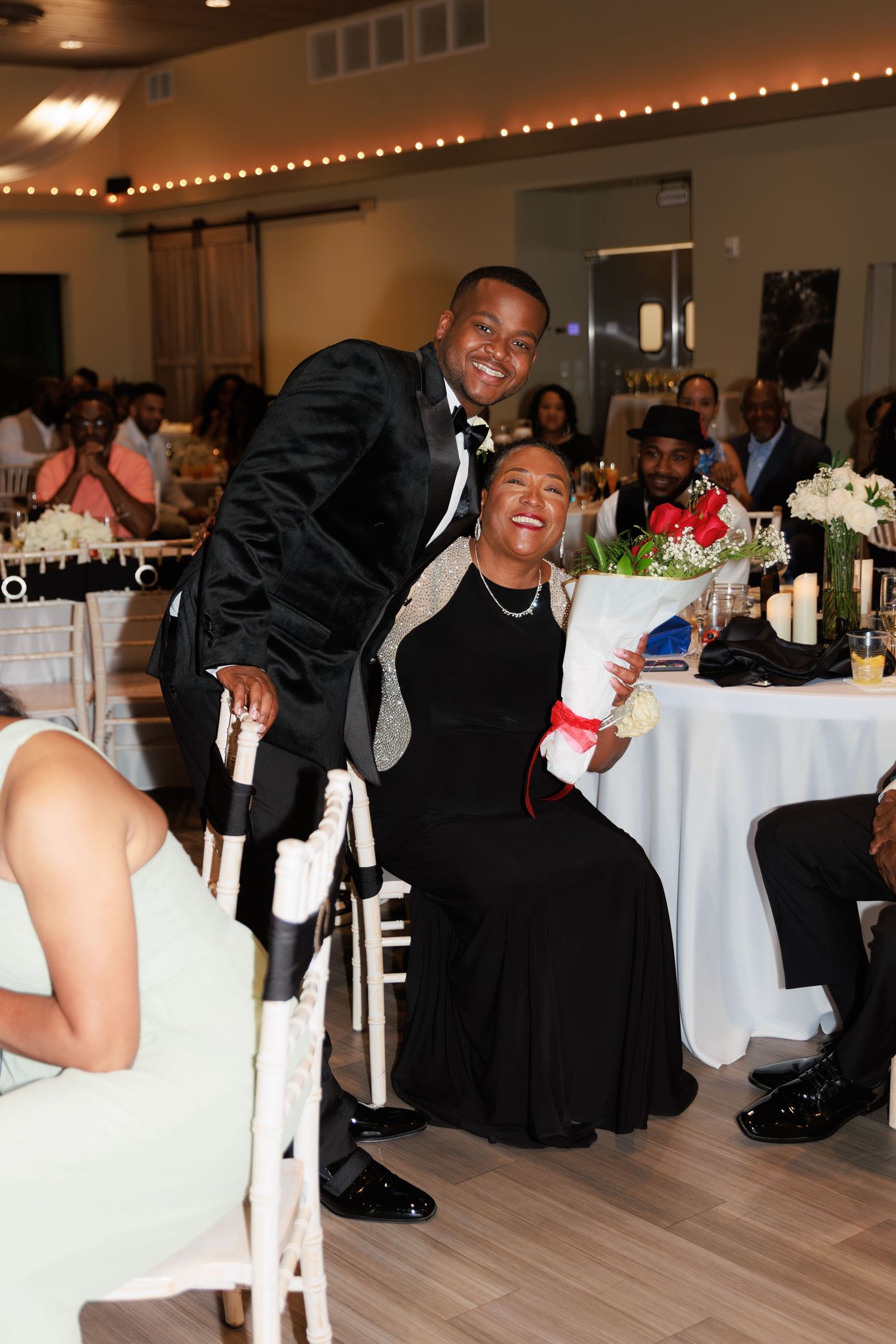 Wedding guests dancing and laughing on the dance floor at an indoor reception with white tablecloths and string lights.