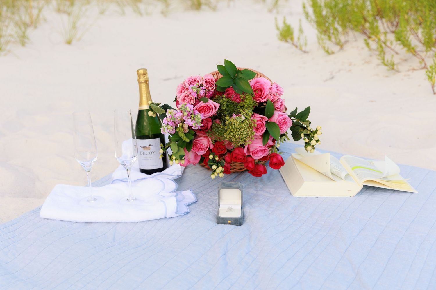 A romantic beach picnic setup with champagne bottle, pink floral bouquet, and white linens on sand.
