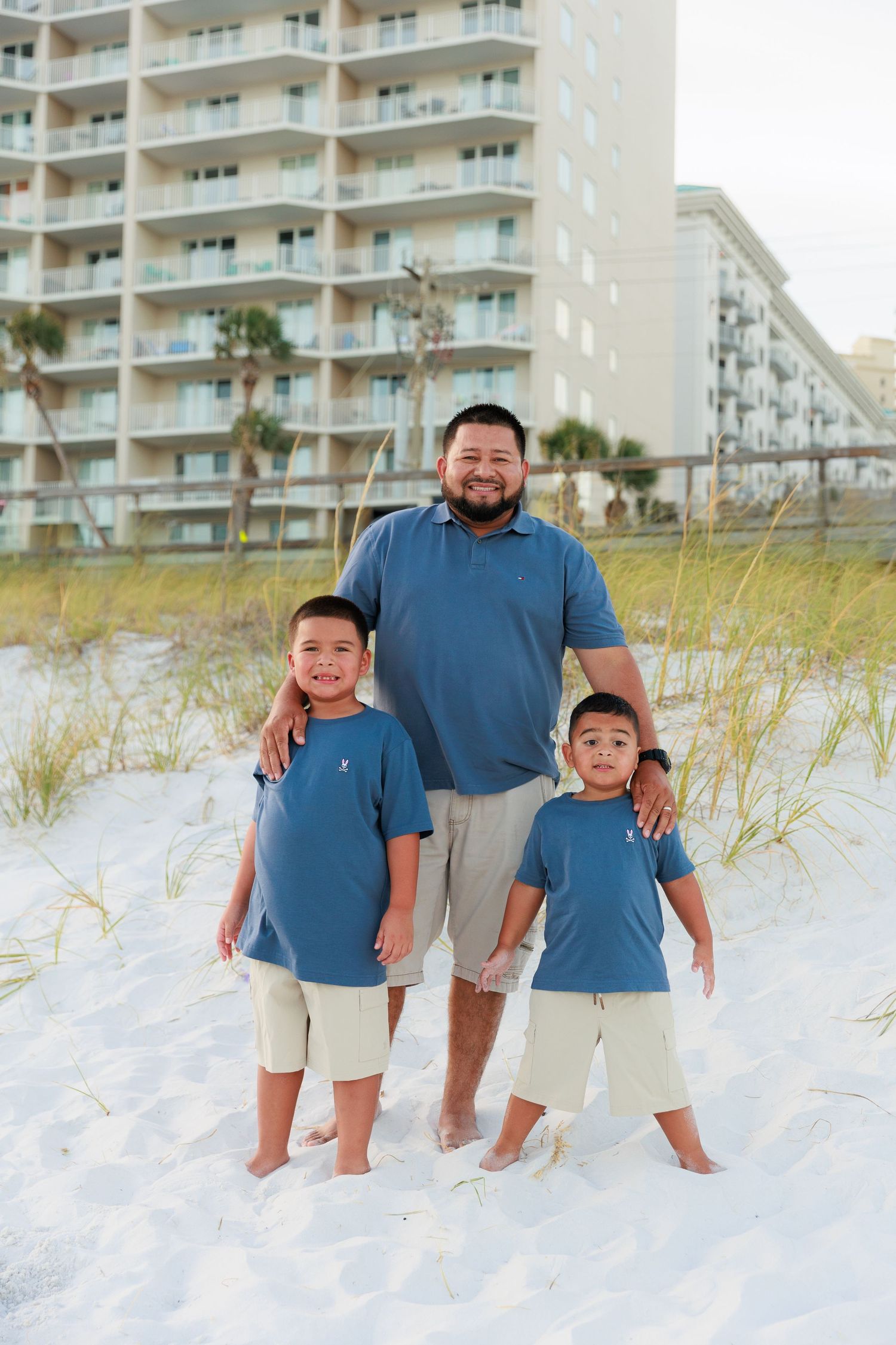 A family poses together on a white sandy beach with beachfront condos in the background.