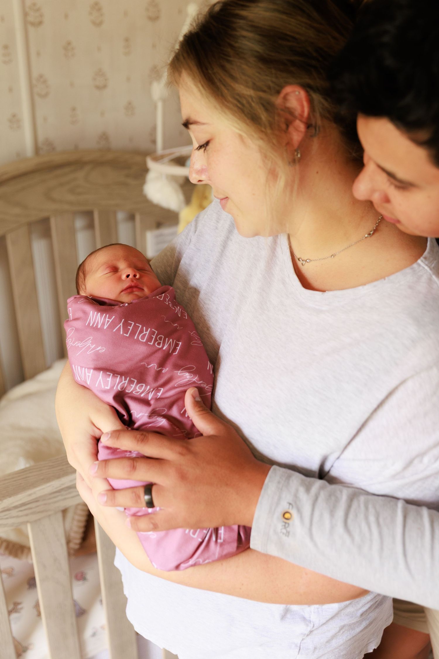 Parents in white shirts tenderly hold and gaze at their newborn baby wrapped in pink blanket in nursery setting.