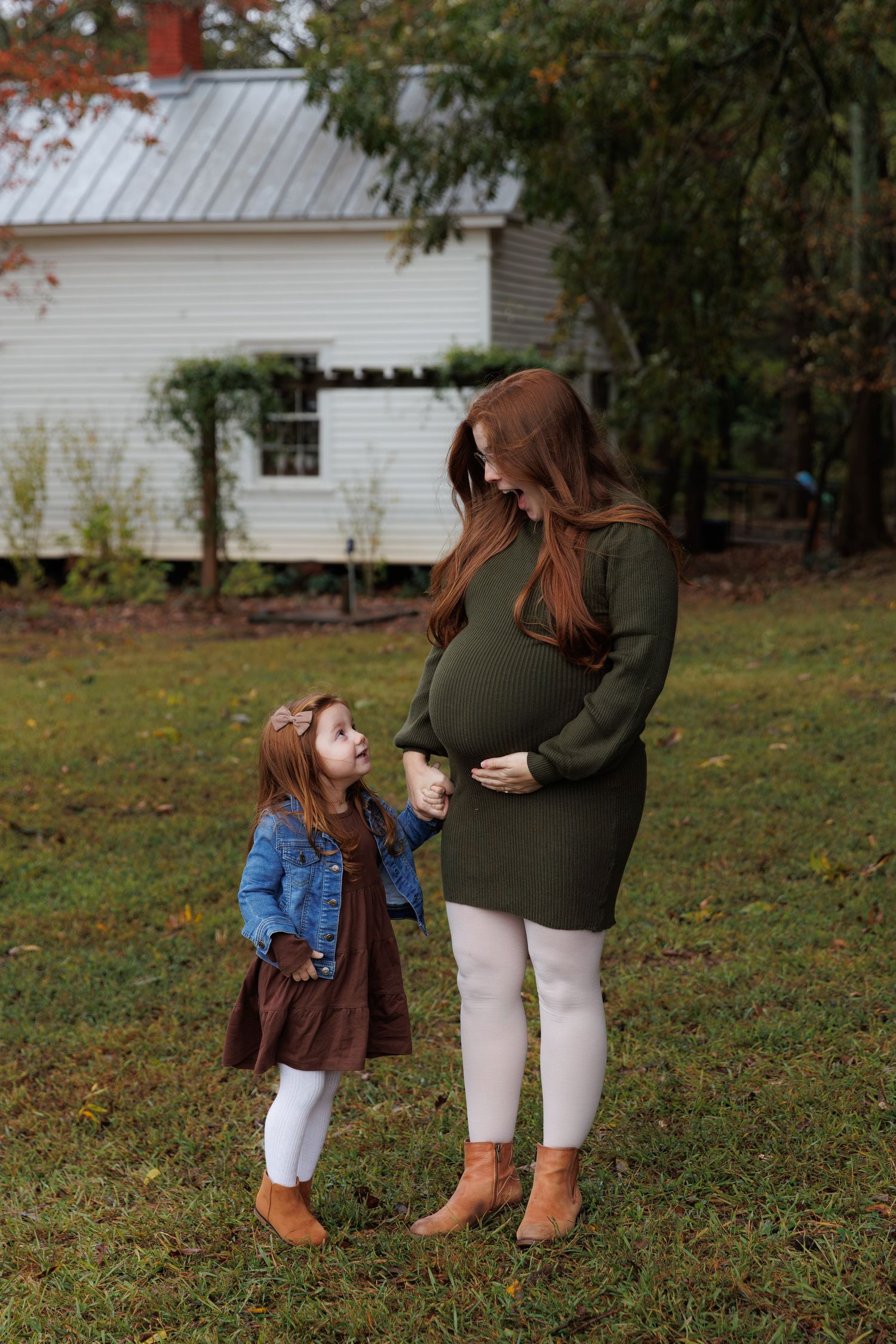 A parent and child wearing fall outfits share a tender moment while standing together on grass near white buildings.
