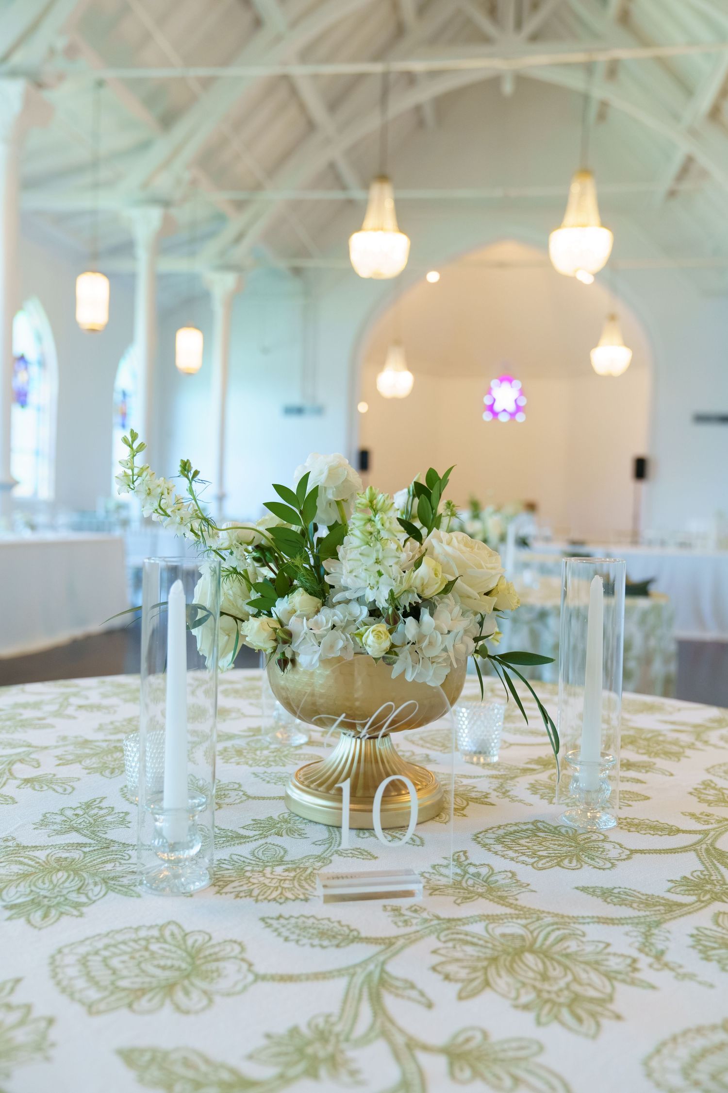 Elegant wedding reception table with gold vase of white flowers, green floral tablecloth, glass candleholders, and table number 10.