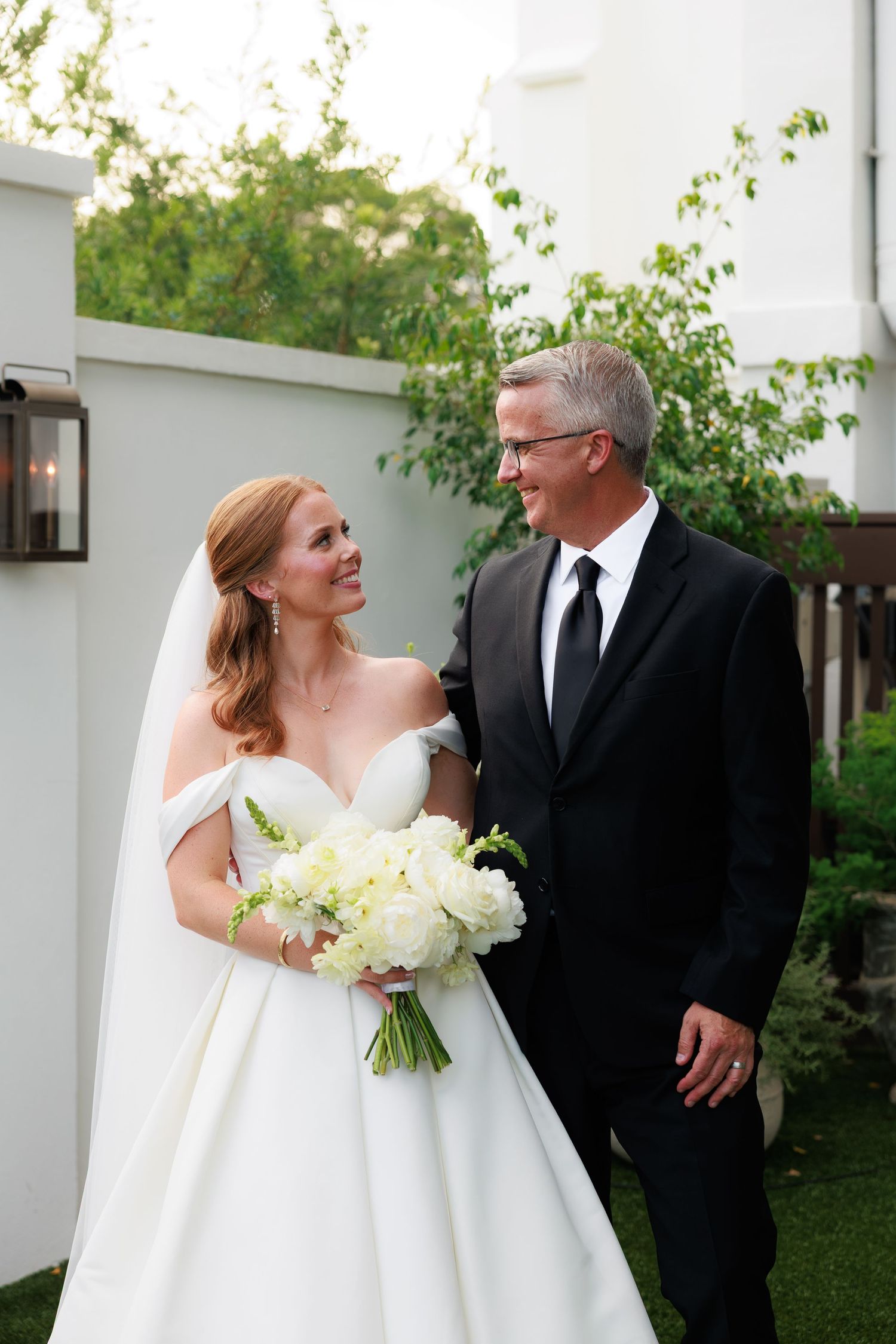 Bride in off-shoulder white gown holding bouquet smiles at groom in black suit outdoors at wedding.