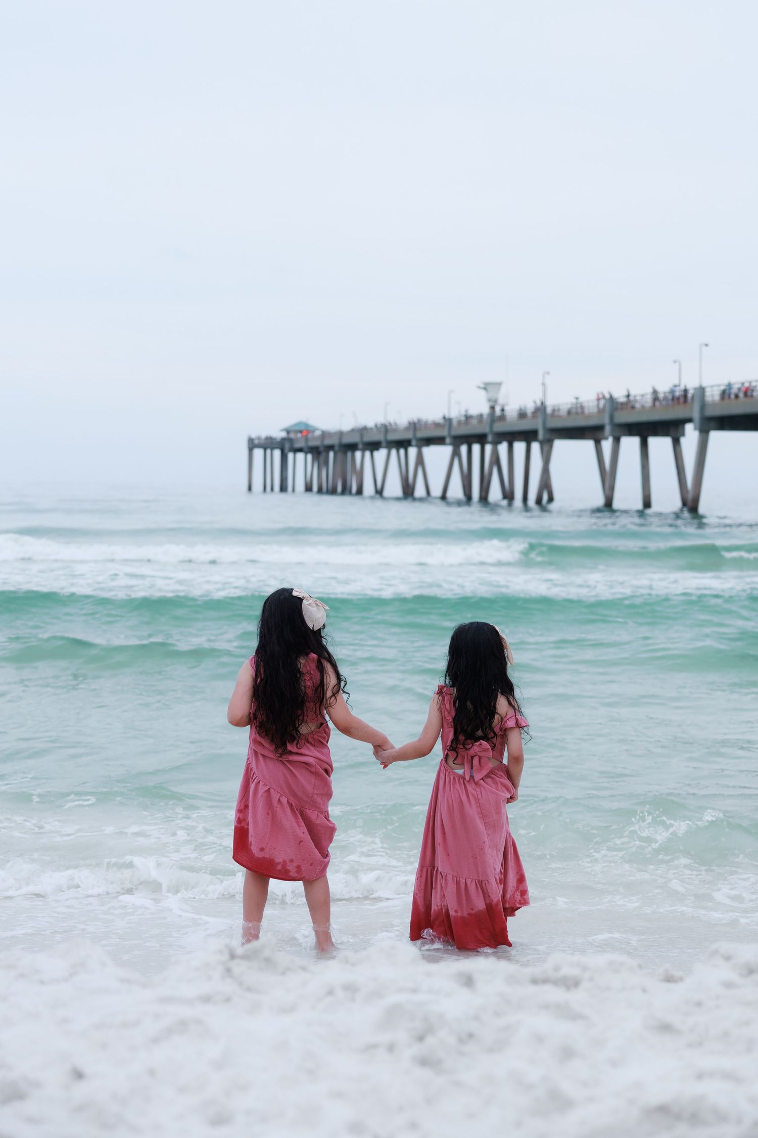 Two sisters in matching pink dresses holding hands facing ocean waves near a beach pier on overcast day.