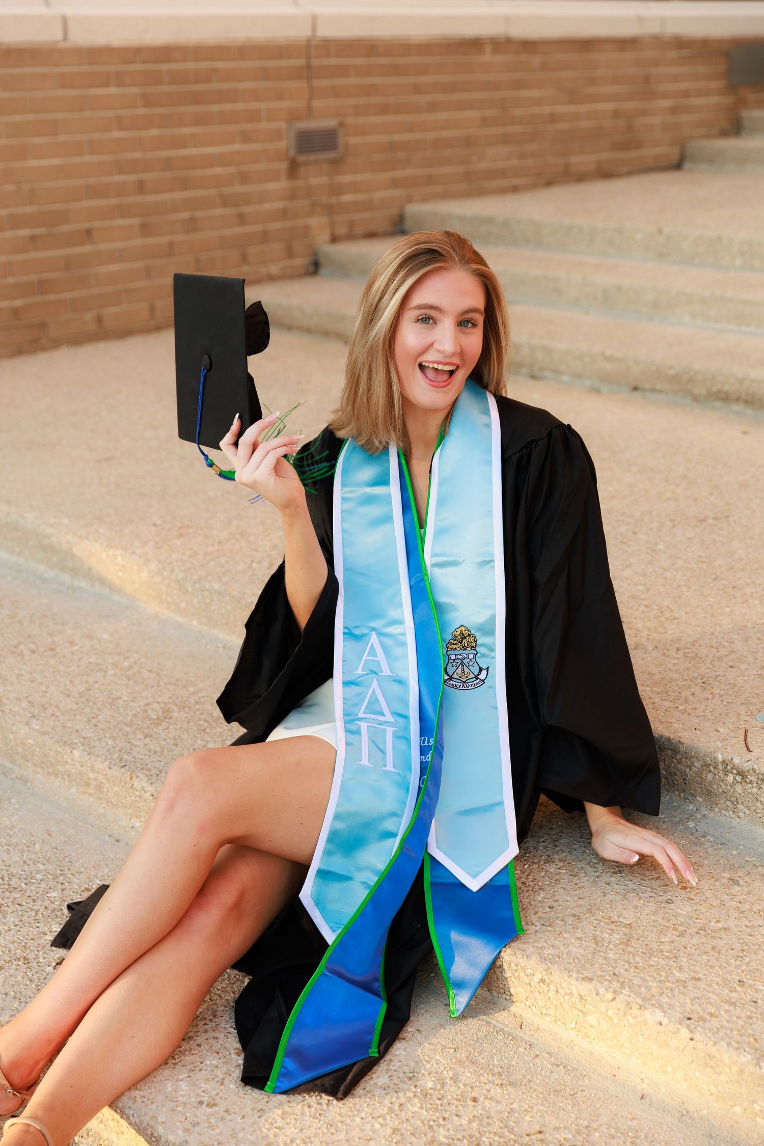 Graduate in black cap and gown with light blue stole sits joyfully on concrete steps holding diploma.