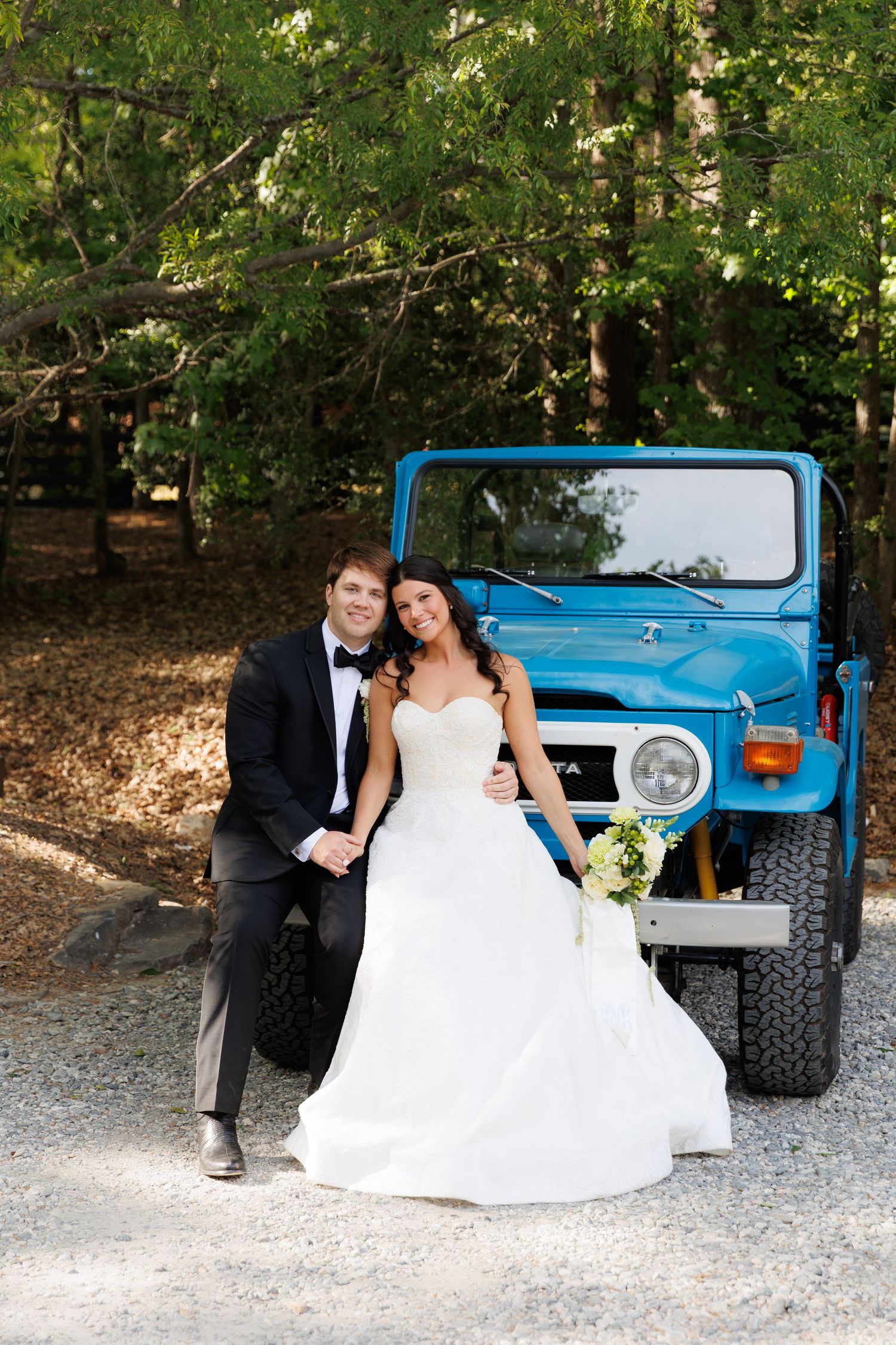 Bride and groom lean against vintage blue Toyota FJ40 in wooded setting, bride holding white floral bouquet.