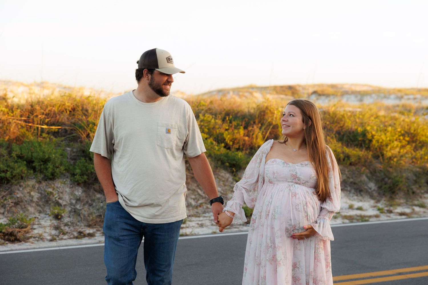 Pregnant couple holding hands on a road at sunset, woman in floral dress cradling baby bump, man in cap and jeans.