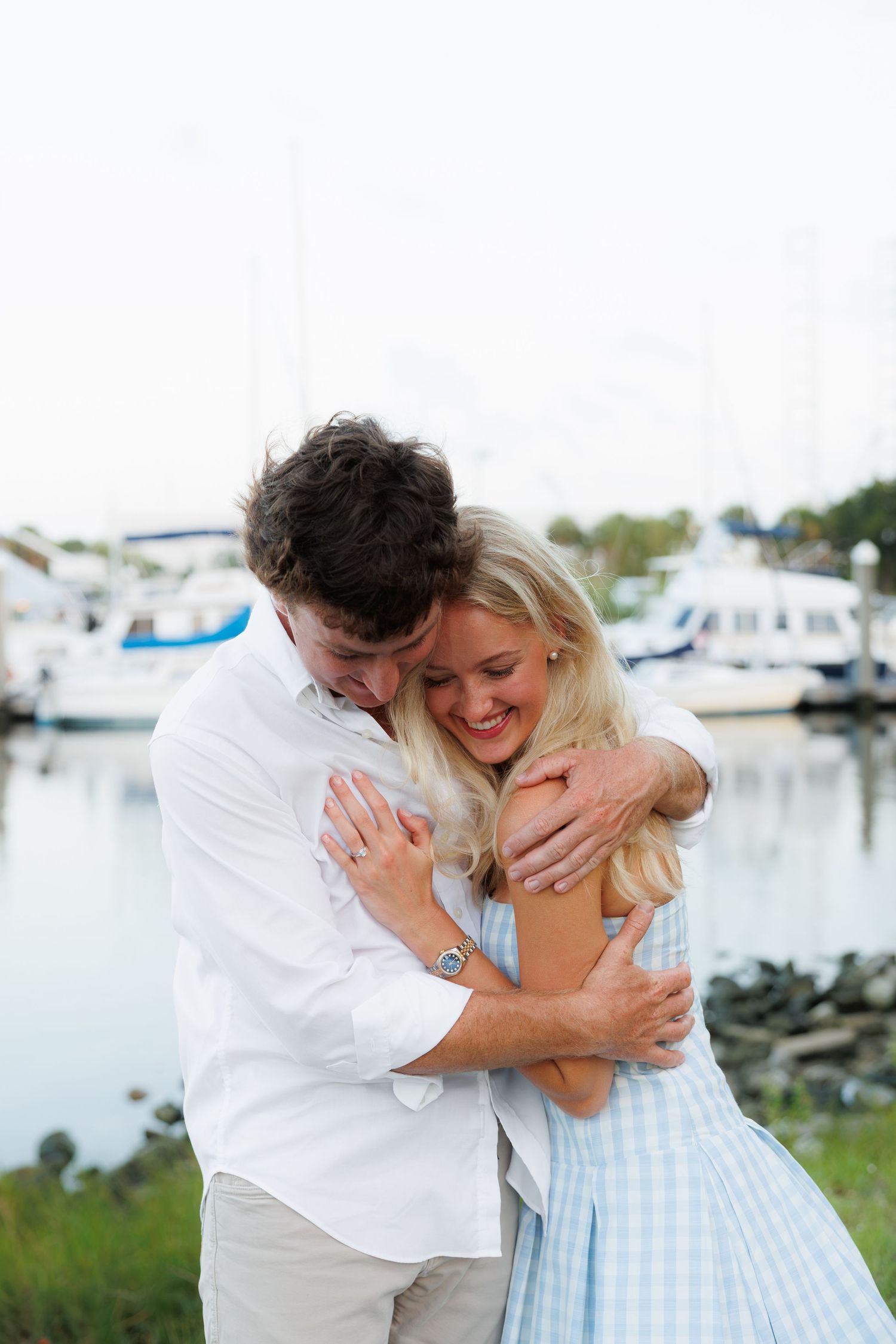 A couple embracing and laughing together near a marina, man in white shirt hugging woman in blue gingham dress.