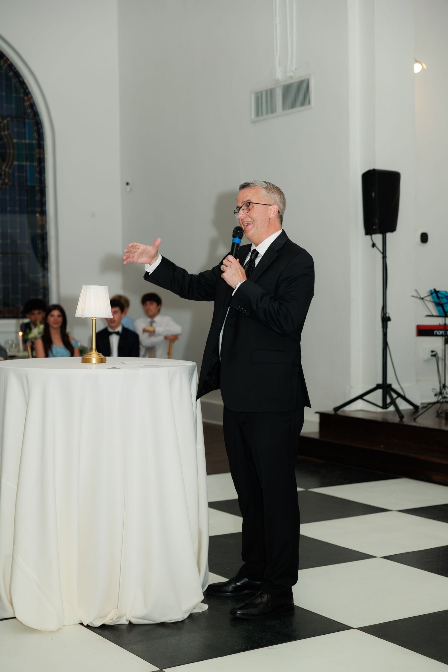 Man in black suit giving a speech at an elegant wedding reception on a checkered dance floor.