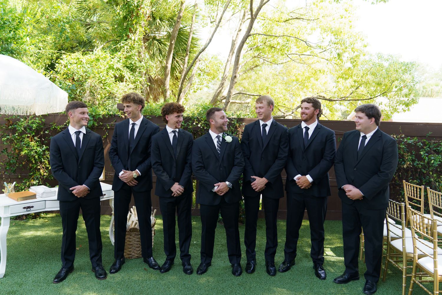 Seven men in matching black suits smiling together outdoors at a wedding ceremony on a green lawn.