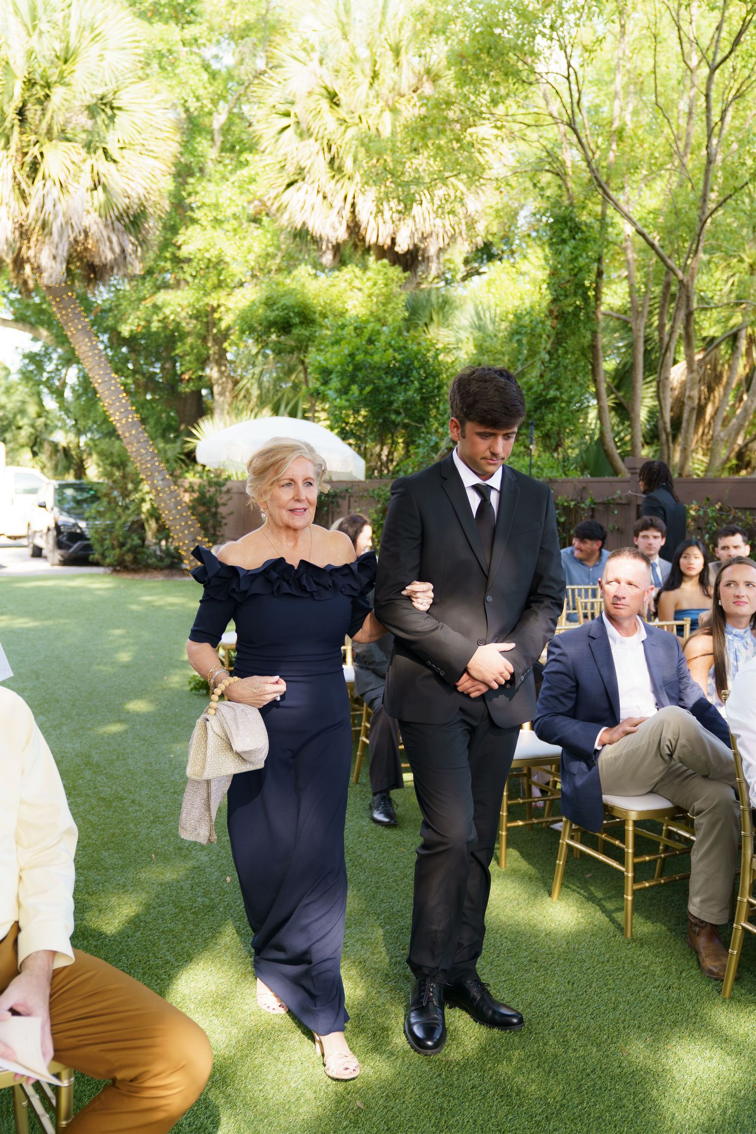 Elderly woman in navy gown escorted by young man in black suit down aisle.