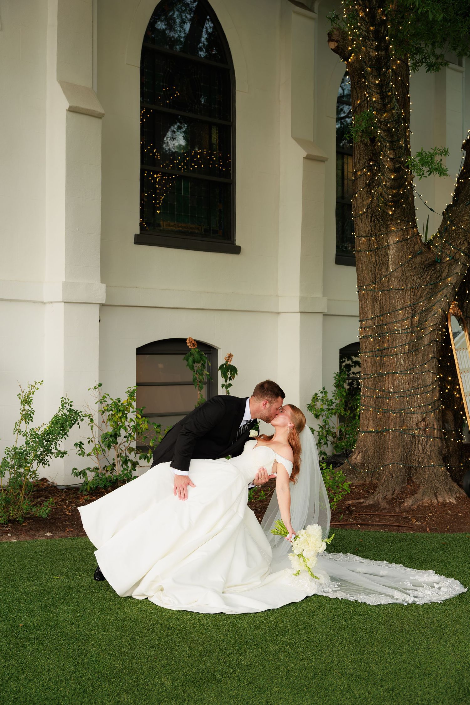 Bride and groom share a romantic dip kiss beside a tree with fairy lights outside a white wedding venue.
