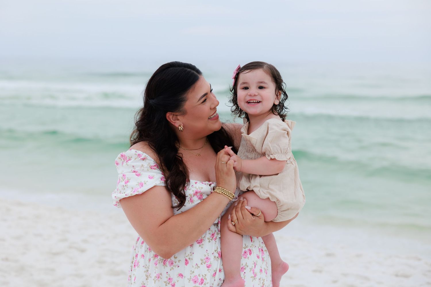 A smiling mother holds her happy toddler daughter on a sandy beach with turquoise ocean waves in the background.