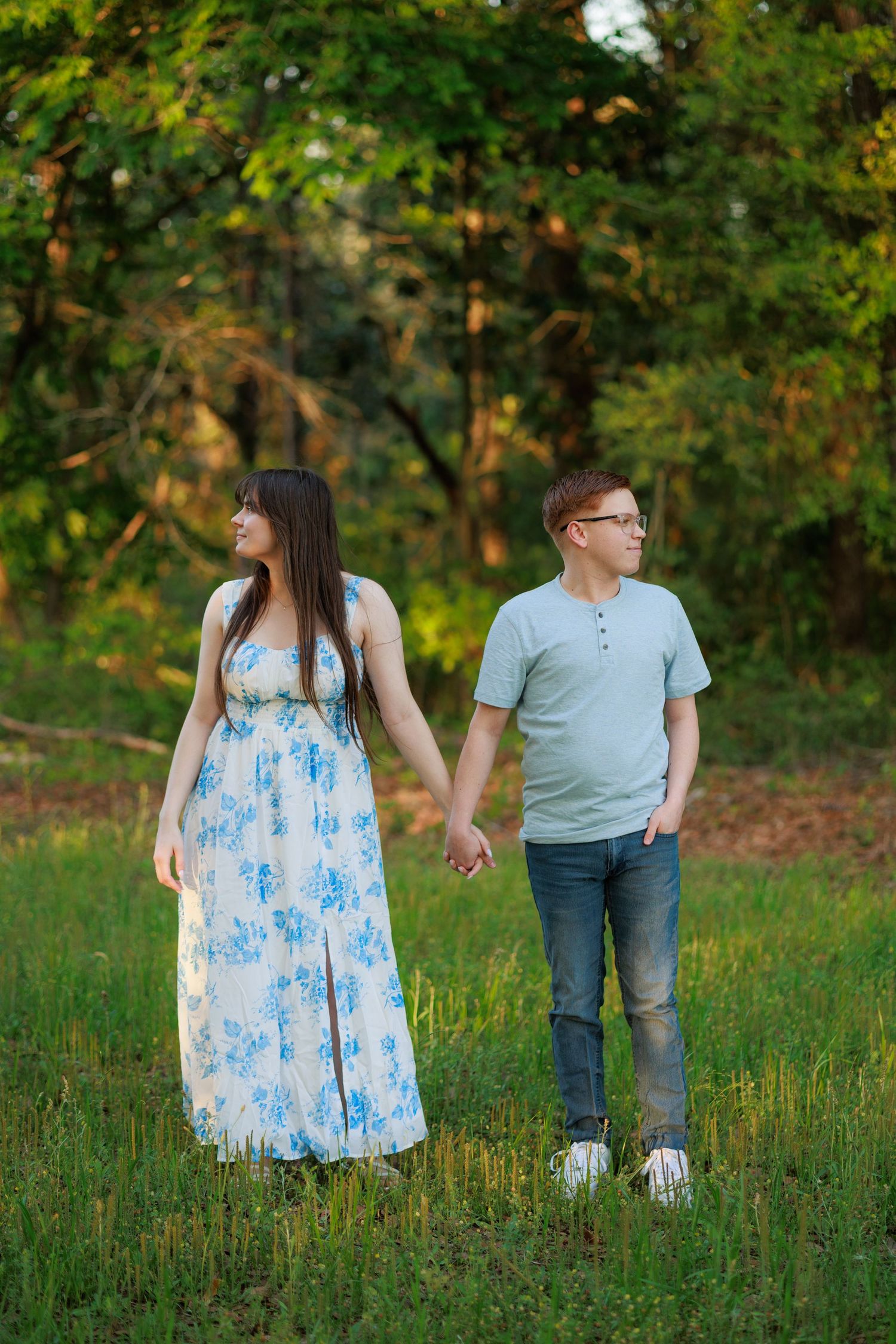Couple holding hands in a green meadow, woman in floral dress and man in casual attire, with lush trees in background.
