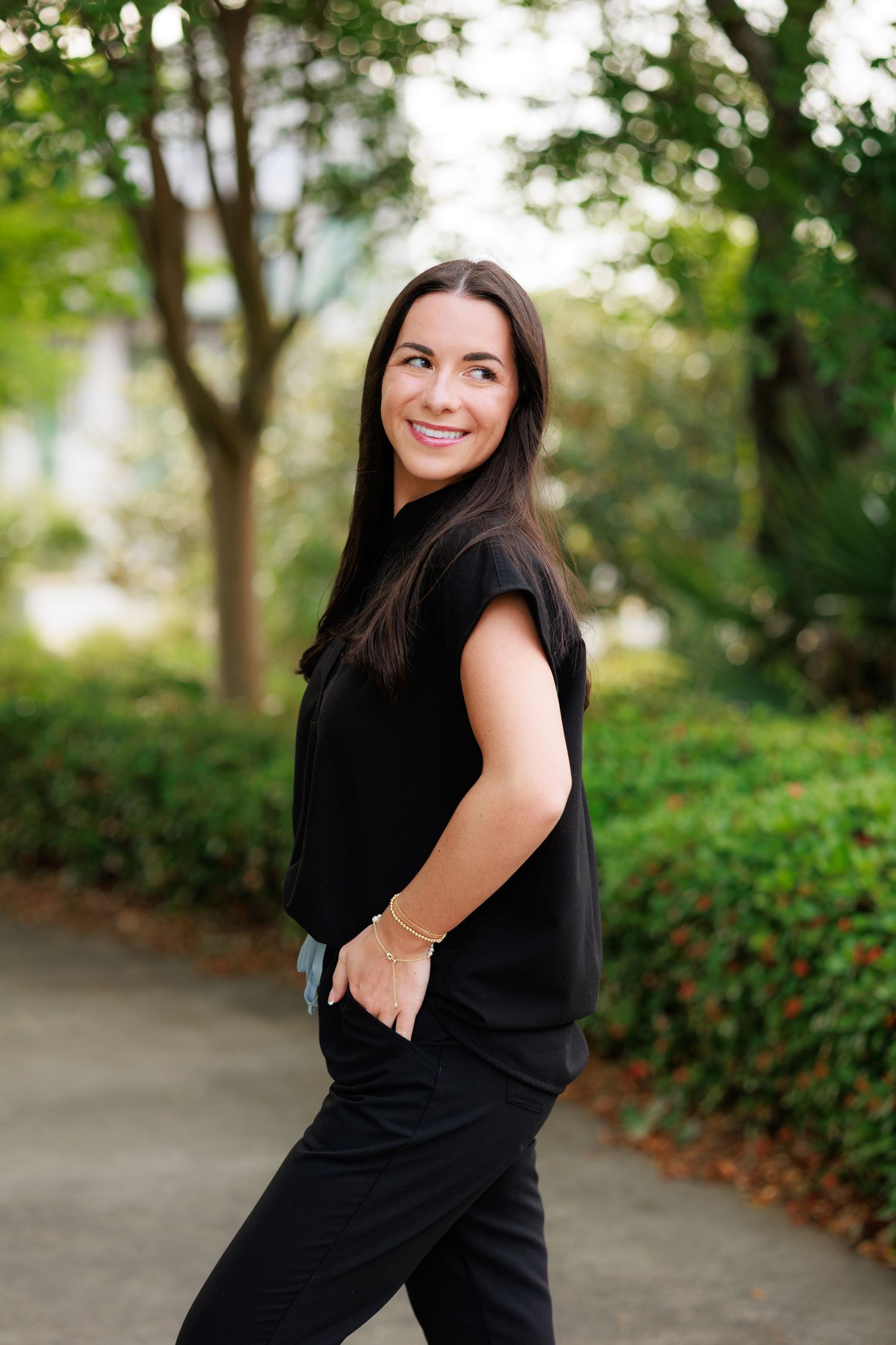 Woman in black outfit smiling over shoulder in lush green park setting, casual outdoor portrait photography.